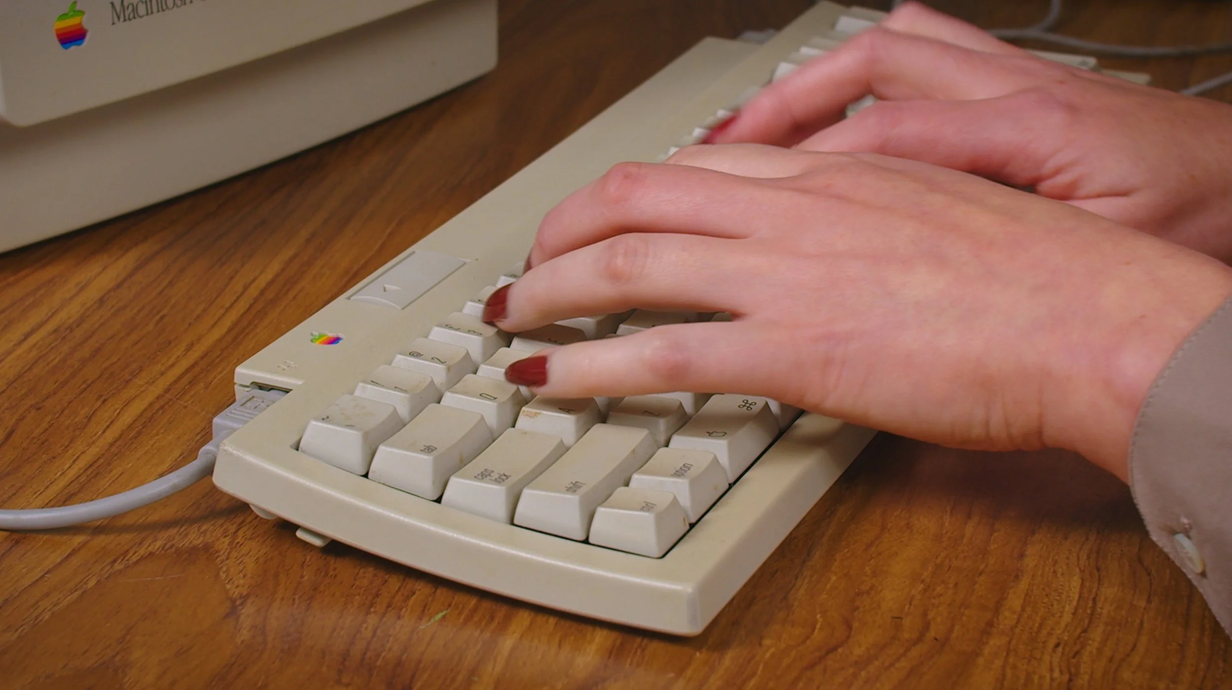 Close-up of a person's hands typing on a vintage beige Macintosh computer keyboard, which has a small rainbow-colored Apple logo on it, placed on a wooden desk.