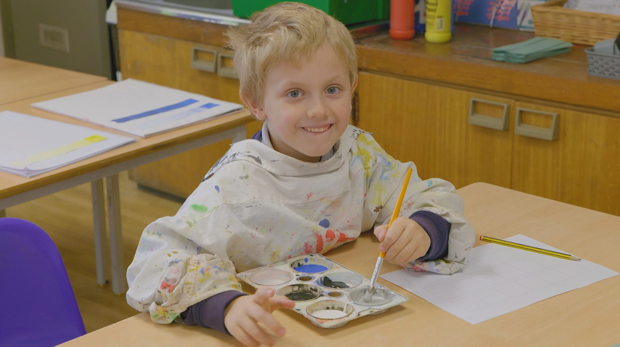 A young boy sitting at a table in a classroom, smiling, holding a paintbrush in one hand and a palette with watercolors, wearing a white smock with colorful paint stains.