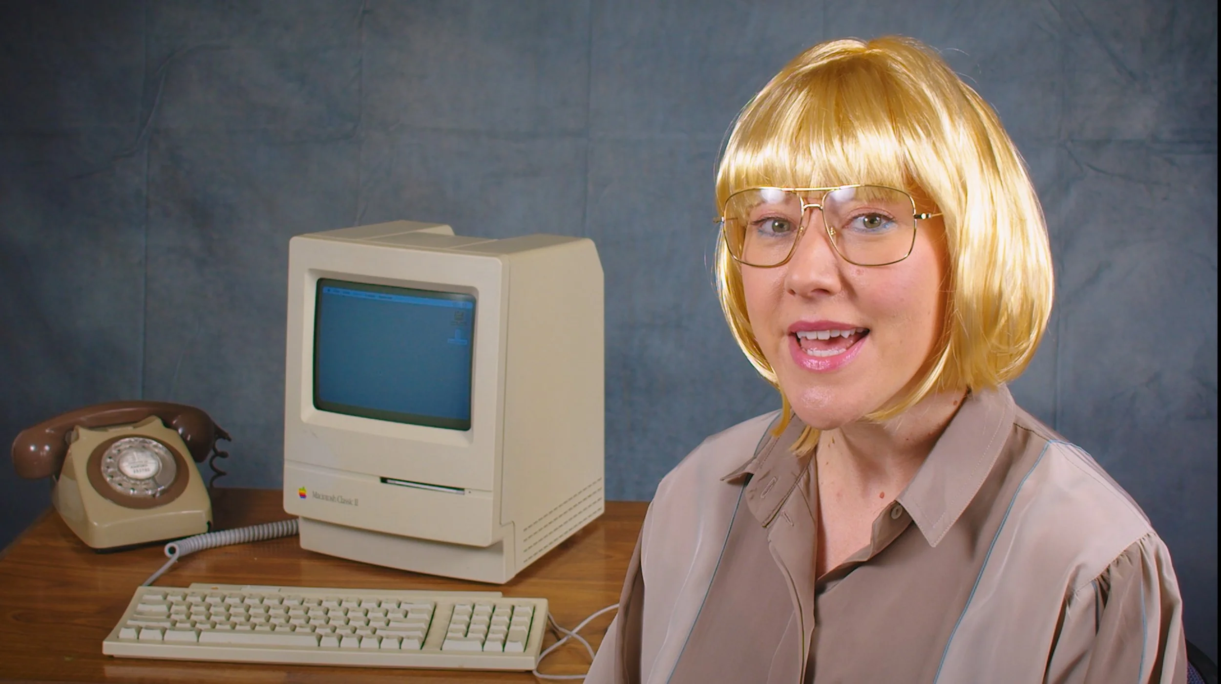A woman with blonde hair and glasses sitting at a desk with an old Apple Macintosh computer, a beige keyboard, and a vintage rotary phone.