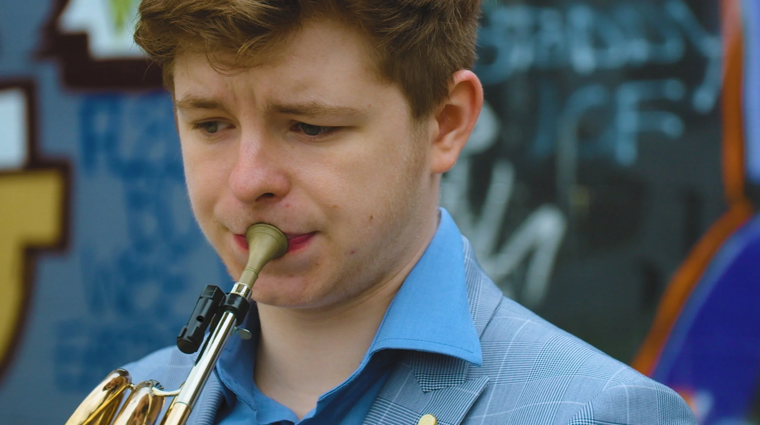 A young man with red hair playing a trumpet, wearing a light blue suit jacket and a blue shirt, in front of colorful blurred background.