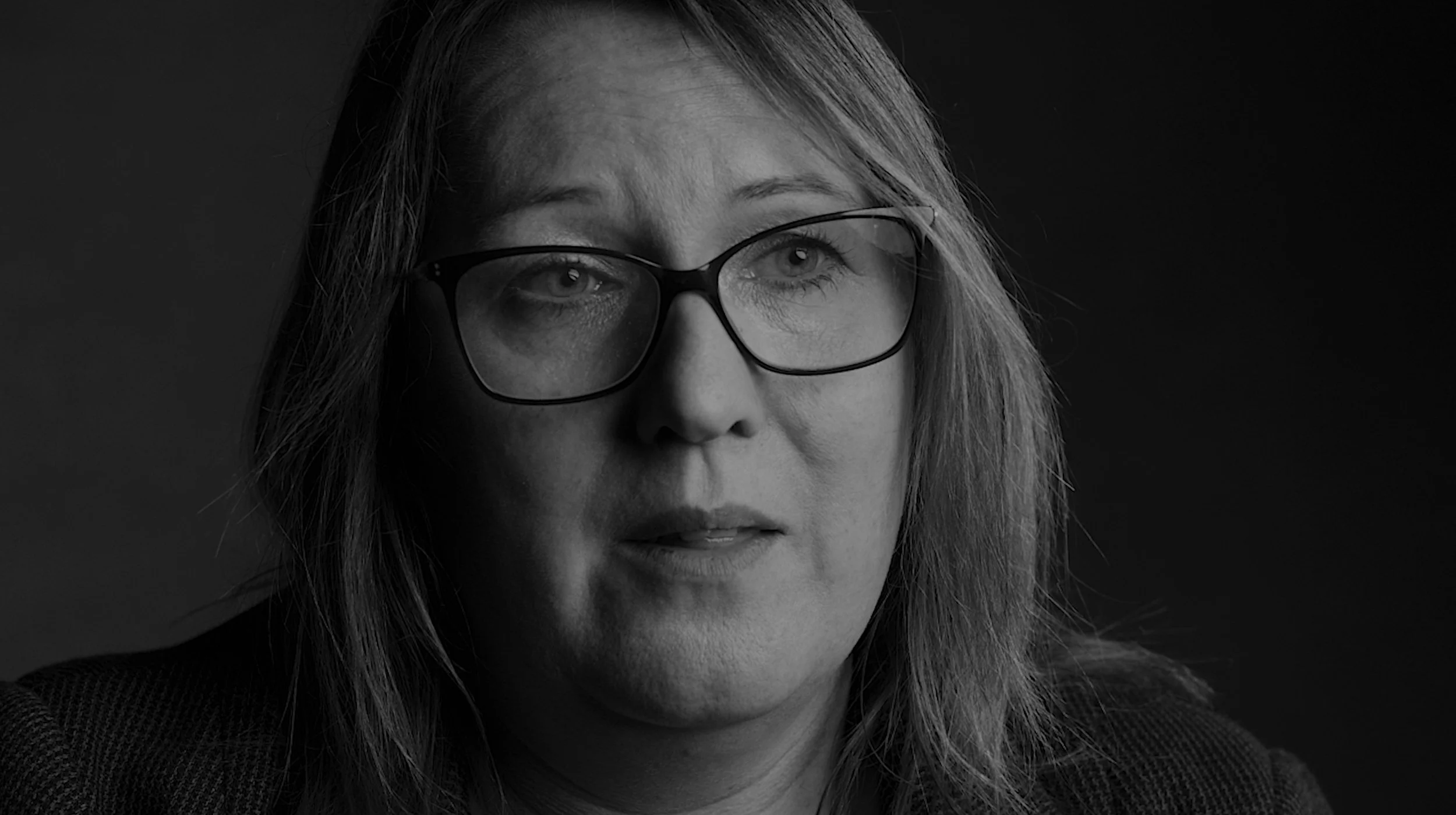 Black and white close-up portrait of a woman with glasses, shoulder-length hair, looking slightly to the side, against a dark background.