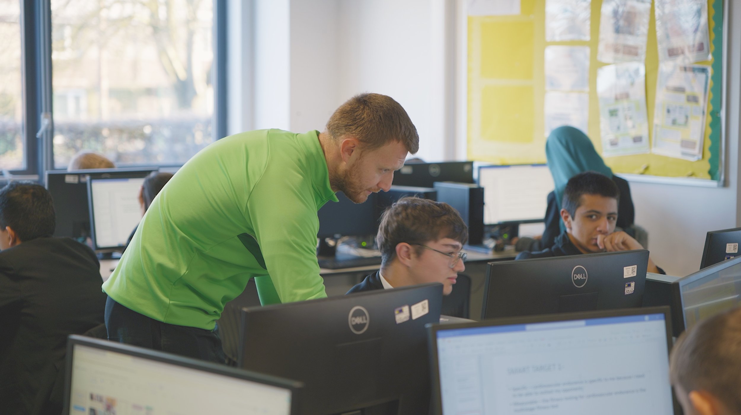 Teacher assisting students in a computer lab classroom with desktop computers.
