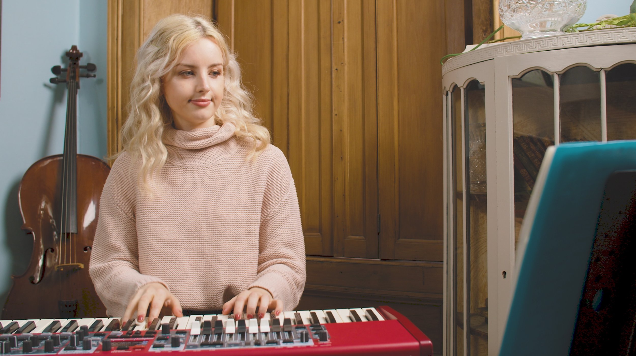 A young woman with blonde curly hair wearing a pink sweater playing a red keyboard in a room with wooden cabinets and a cello in the background.