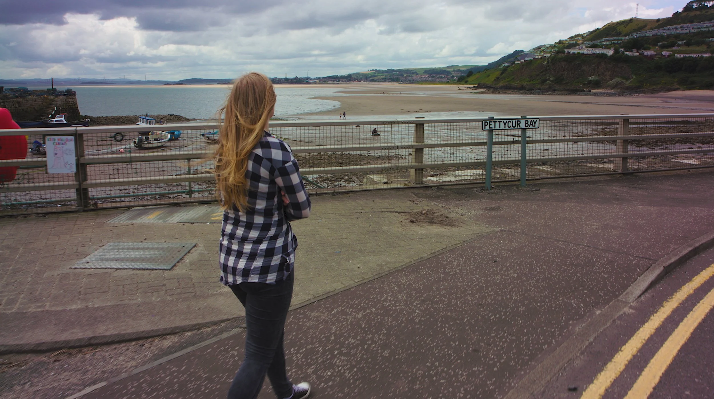 Woman with long hair wearing a checkered shirt standing near a railing overlooking a beach and water, with hills and houses in the background.