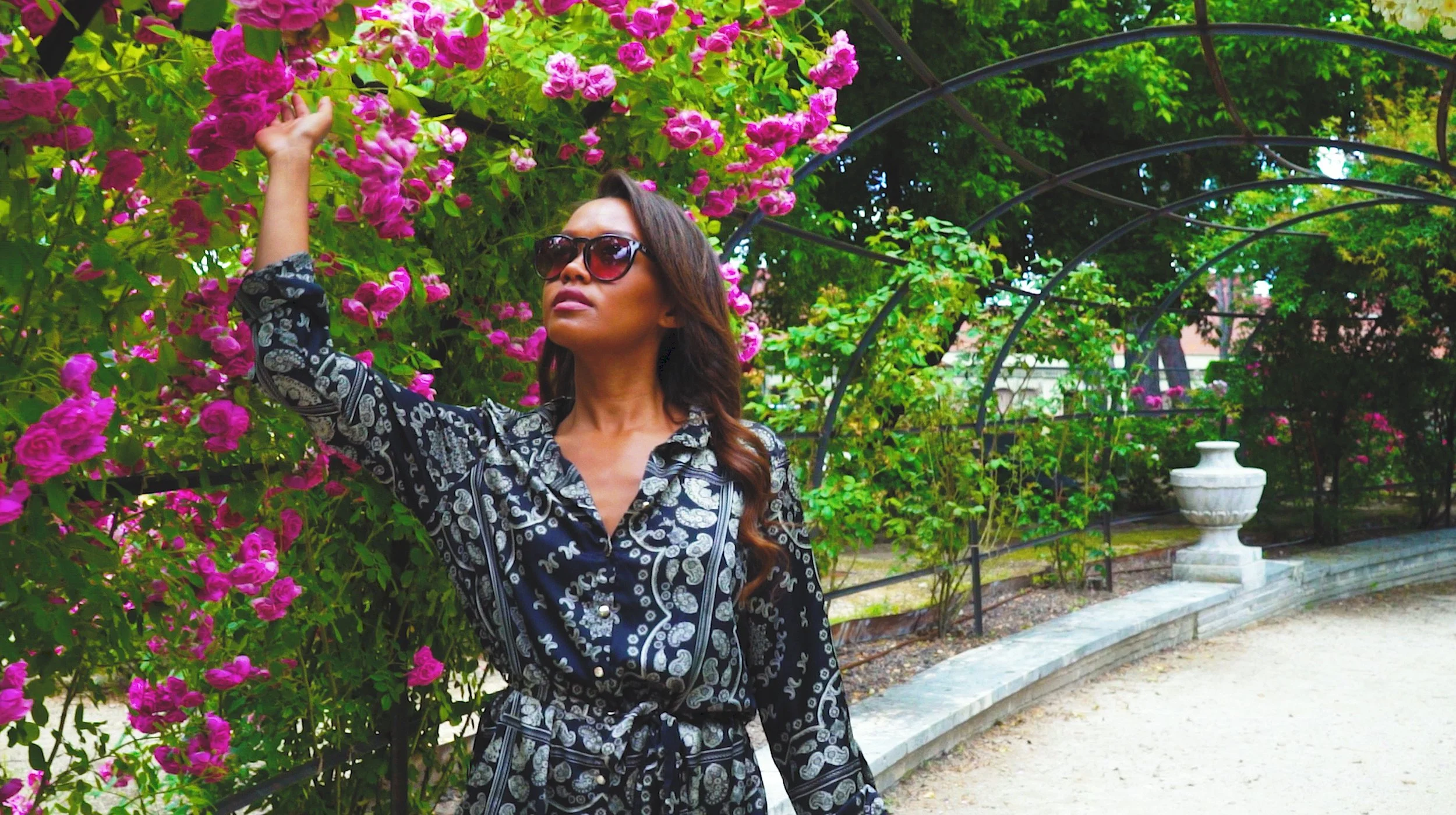 A woman wearing a patterned dress and sunglasses stands in a garden, reaching towards bright pink flowers on an overhead archway.