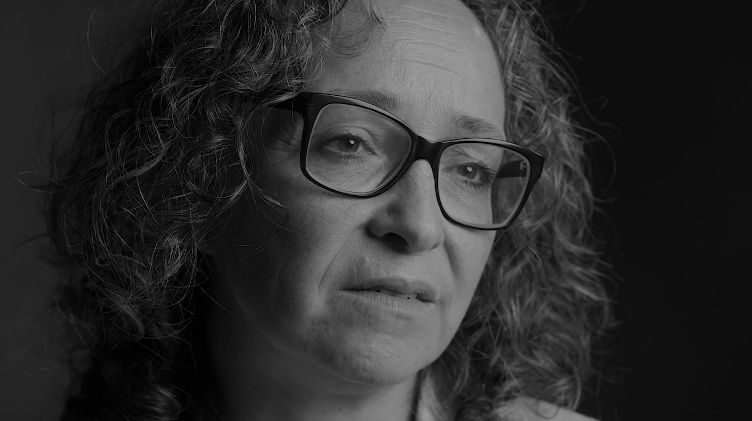 A close-up black and white portrait of a woman with curly hair wearing glasses, looking contemplative.