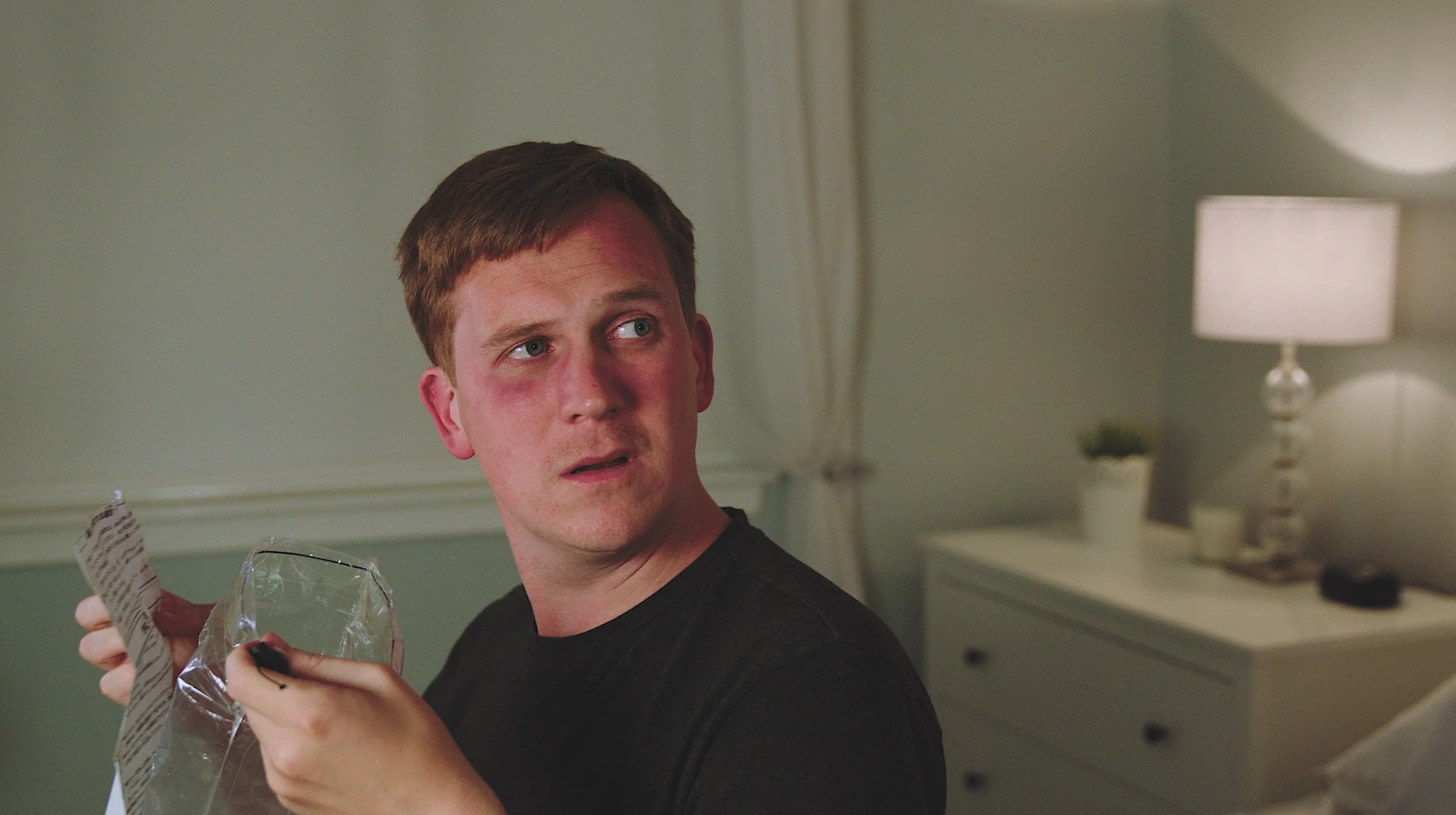 A young man with short brown hair and a reddish complexion looks to the side with a surprised or confused expression while holding open a crumpled plastic bag and some torn paper in a bedroom with a white dresser, a lit lamp, and a small plant in the background.