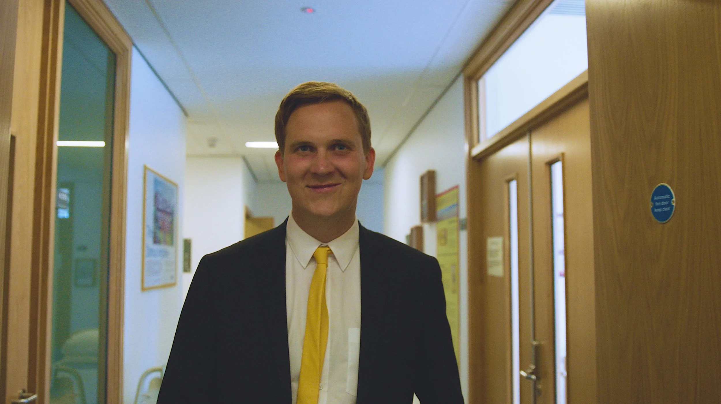 A young man in a suit and yellow tie is standing in a hallway, smiling at the camera.