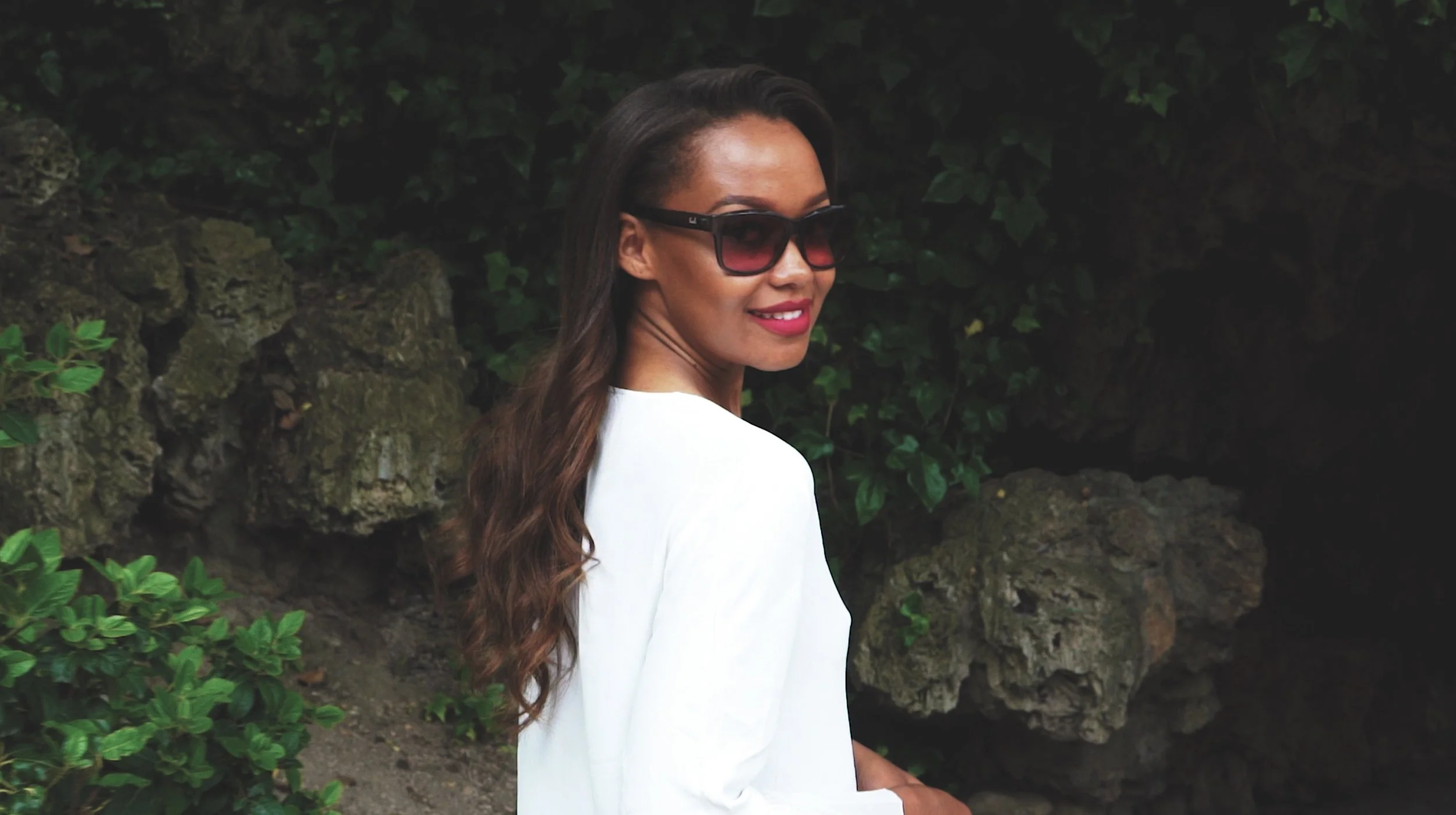 A woman with long wavy brown hair wearing black sunglasses and a white top standing outdoors near green foliage and rocks, smiling at the camera.