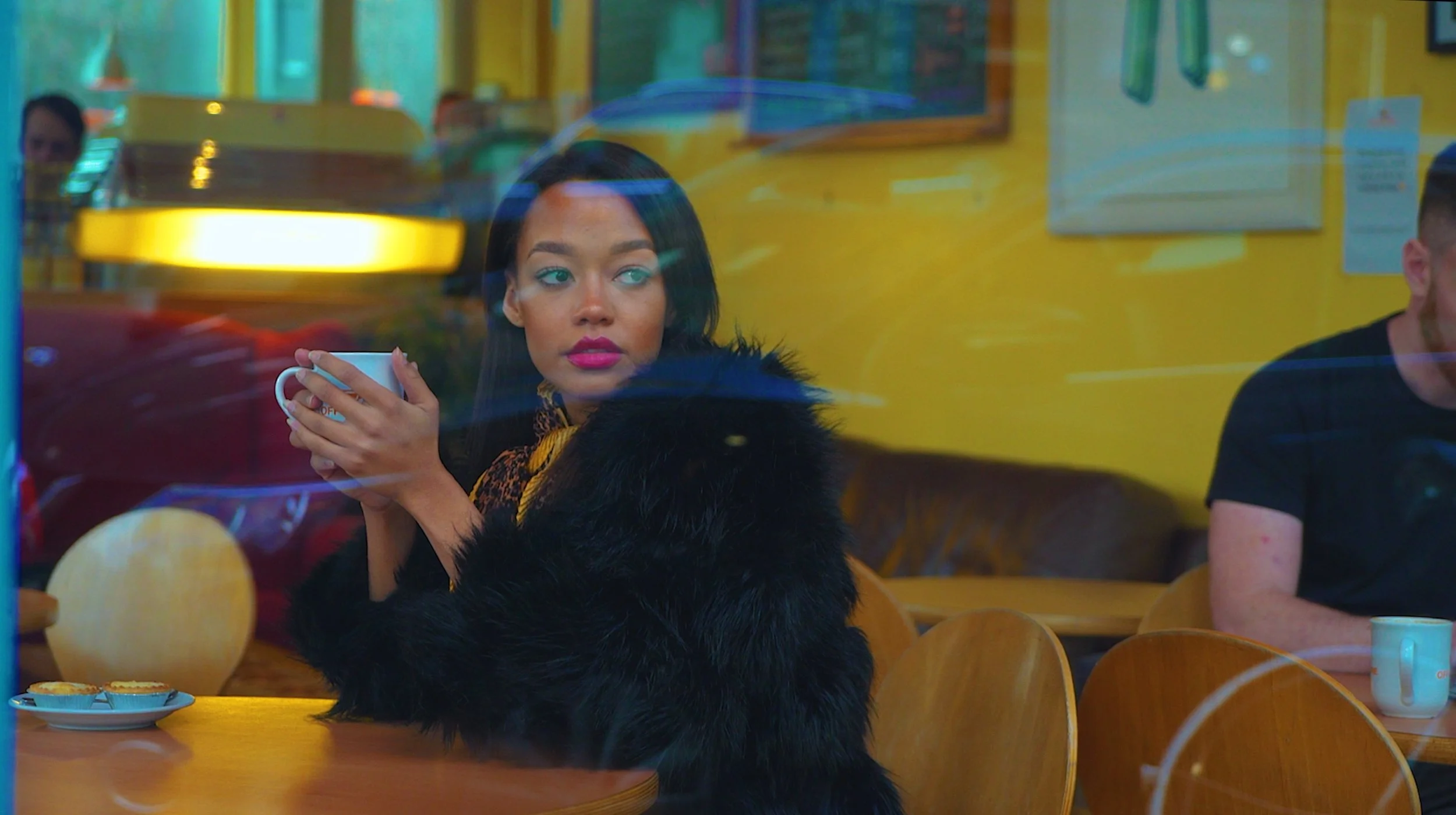 A woman with dark hair and pink lipstick holding a white coffee mug in a colorful cafe with yellow walls and wooden chairs, viewed through a window with reflections.