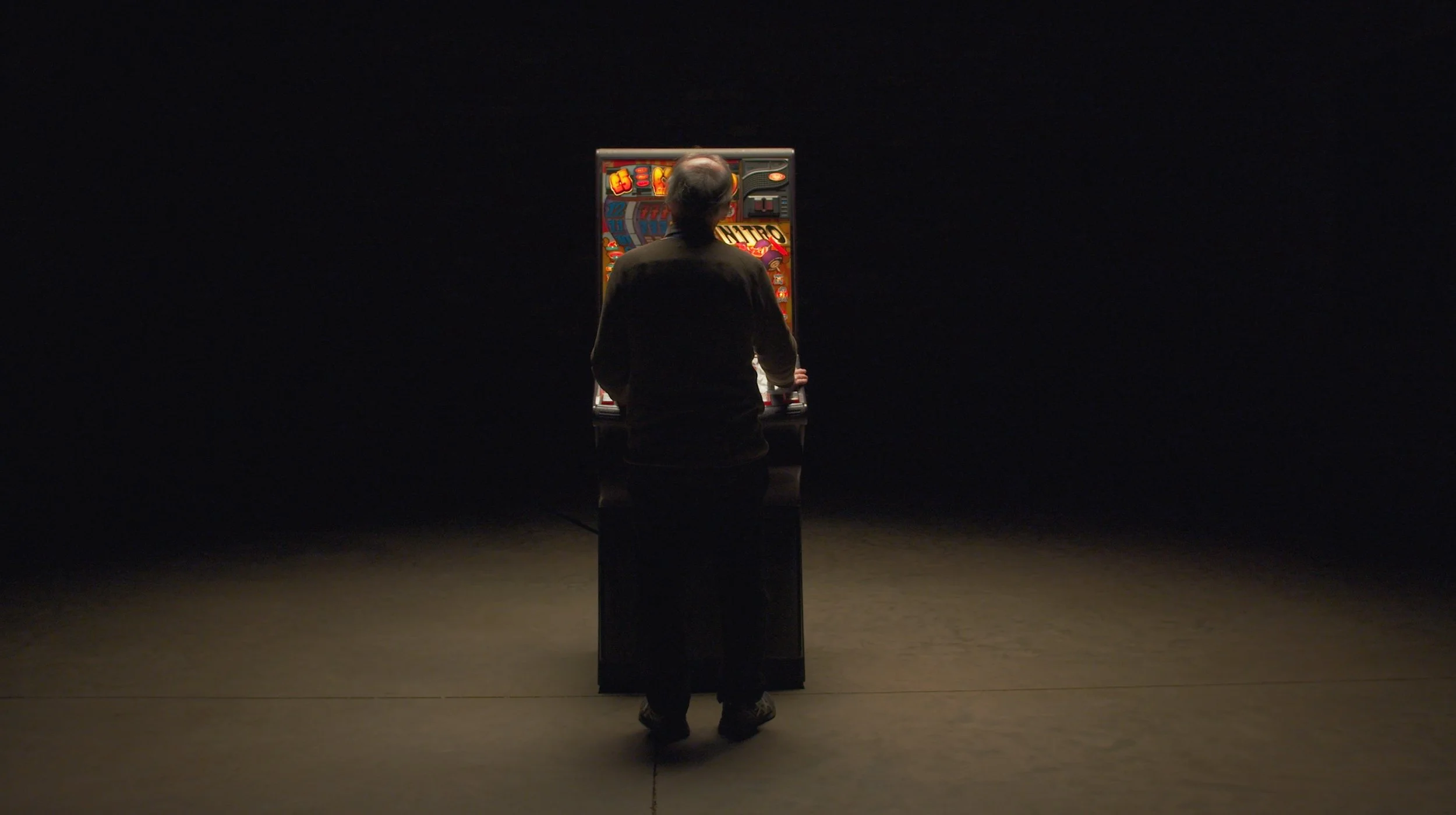 A person standing in a dark room, facing a brightly lit pinball machine on a pedestal.