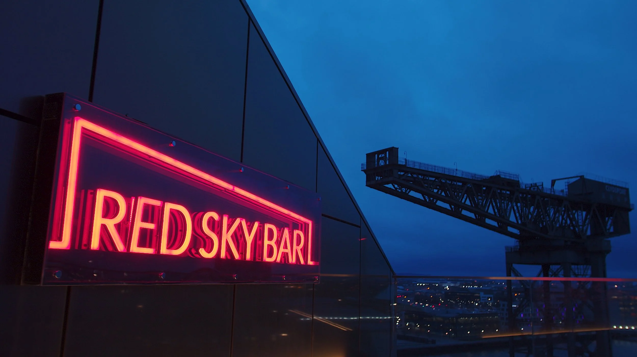 Neon sign reading 'RED SKY BAR' on a dark building wall with city lights and a large crane in the background at dusk.