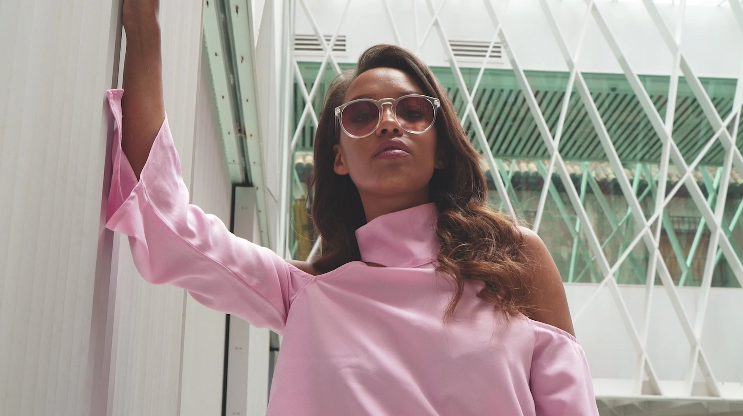 Person wearing sunglasses and a pink off-shoulder top, posing indoors beside a modern architectural wall.