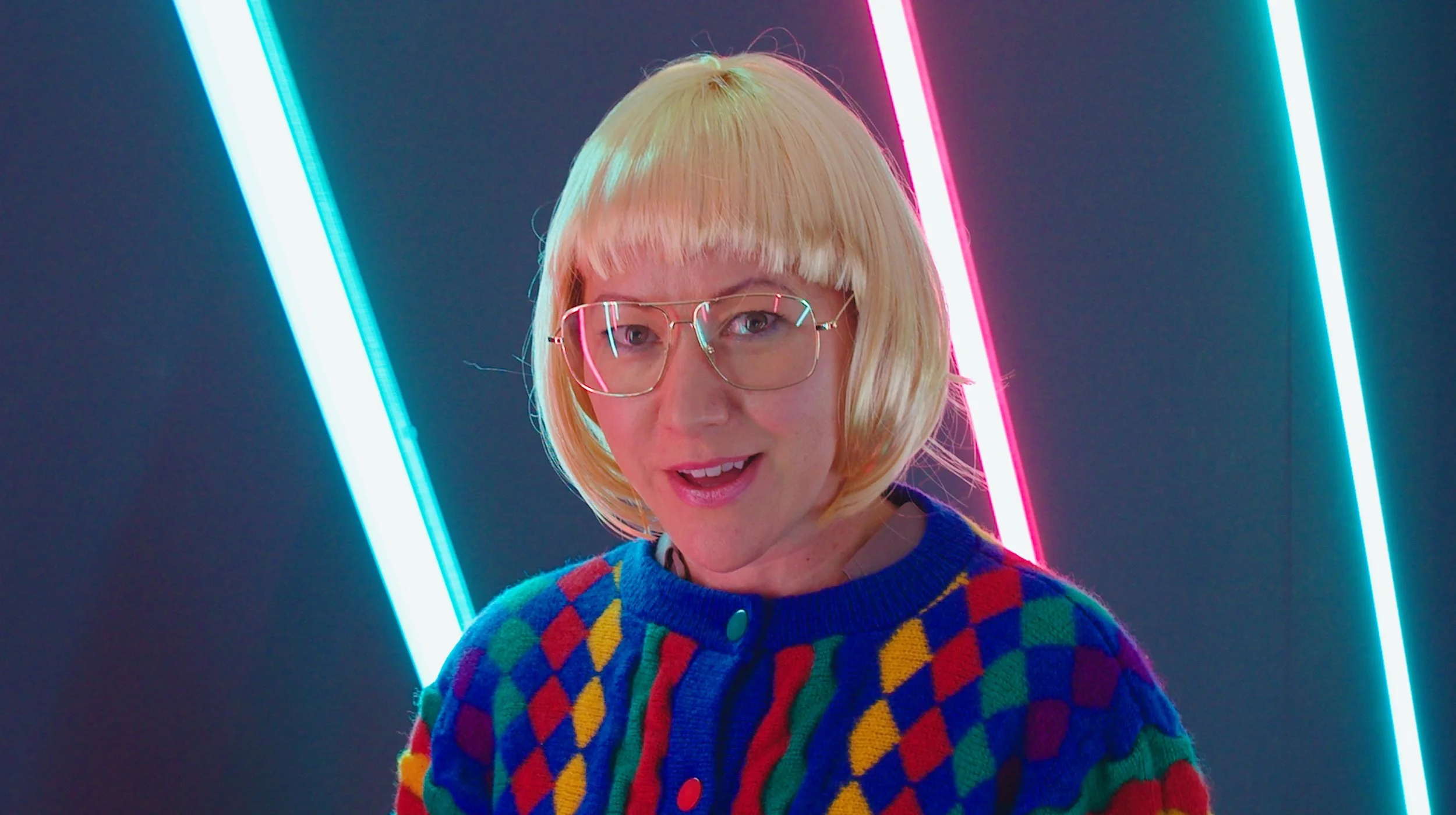 A woman with blonde hair and glasses smiling, wearing a colorful argyle sweater, is standing in front of a dark background with pink and blue neon lights.