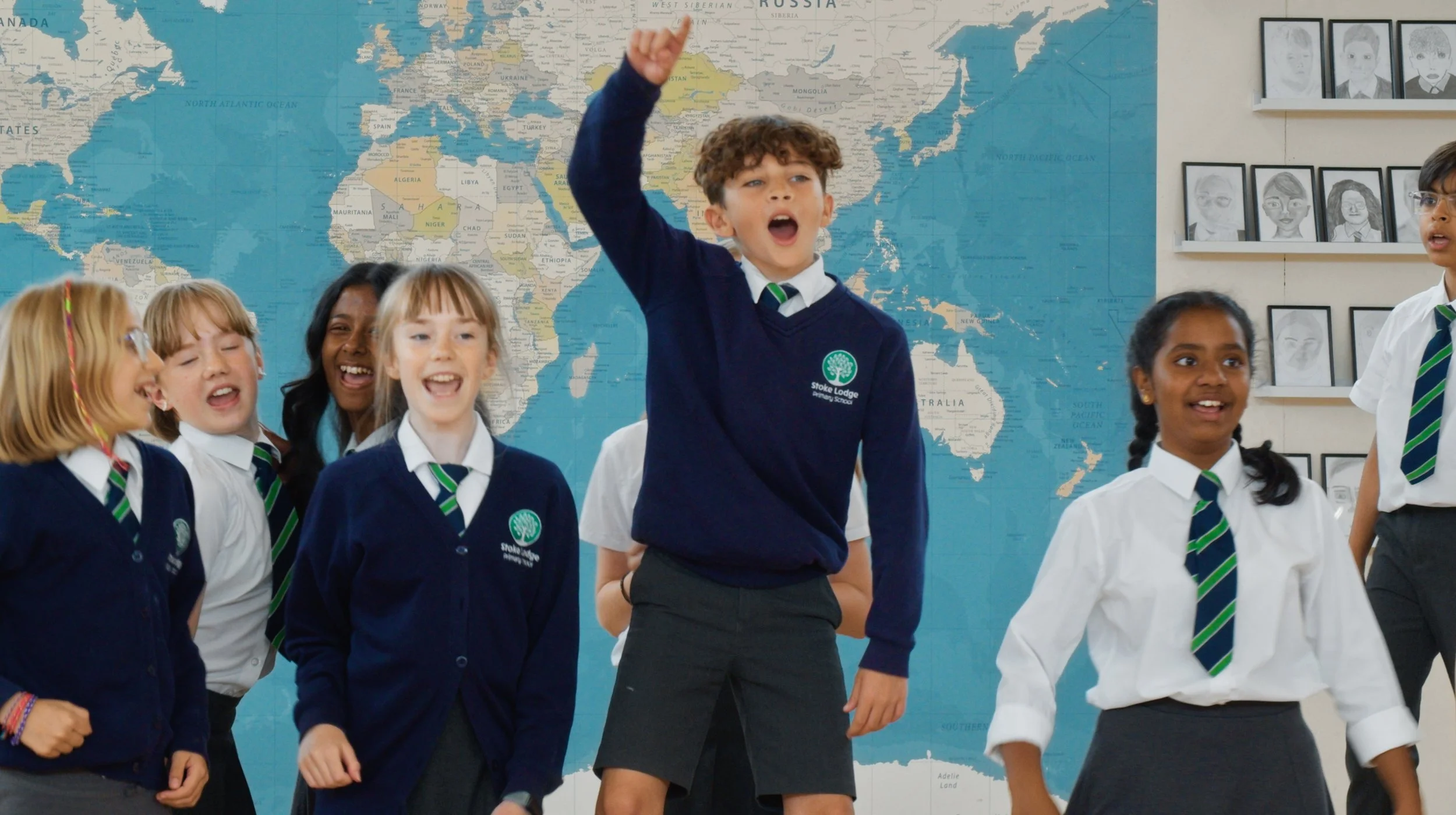 Group of school children in school uniforms, some laughing and smiling, standing in front of a world map on a classroom wall with framed student artwork.