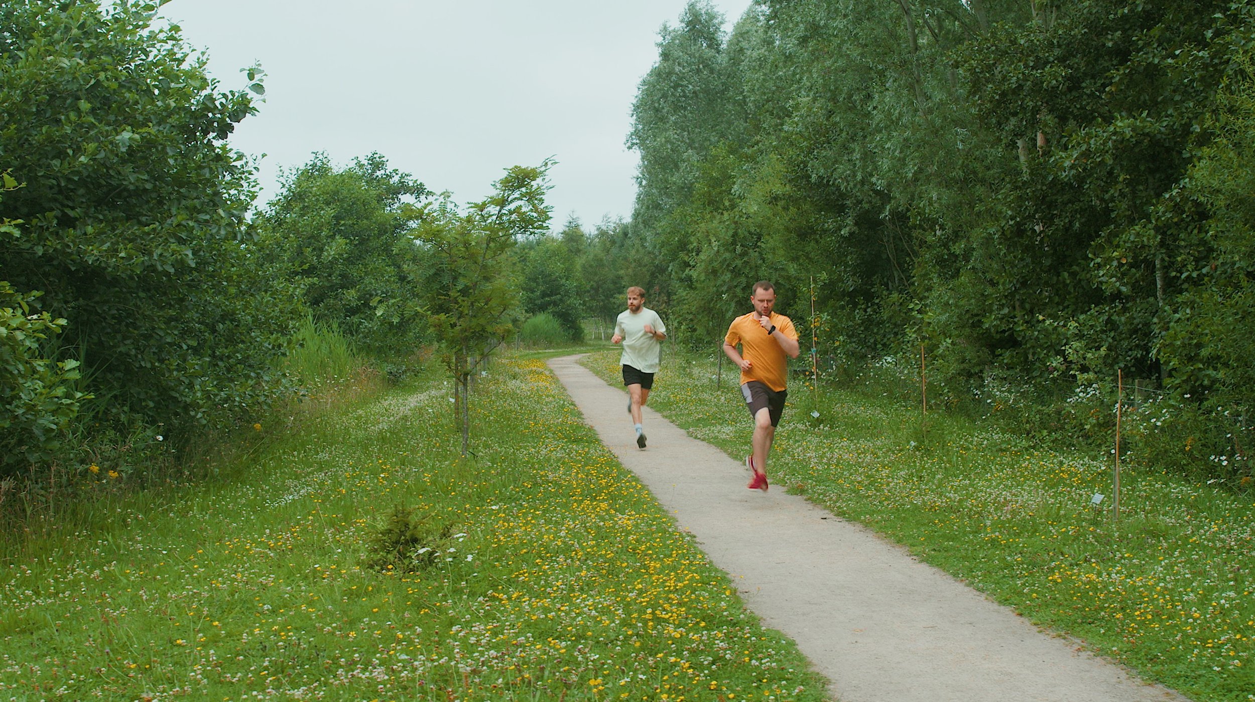 Two men running on a trail in a green park surrounded by trees and grass with wildflowers.