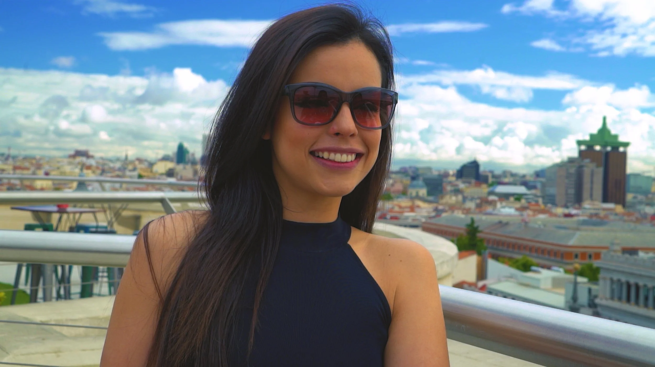 A woman with long dark hair wearing sunglasses and a sleeveless black top, smiling on a rooftop with a city skyline in the background under a partly cloudy sky.