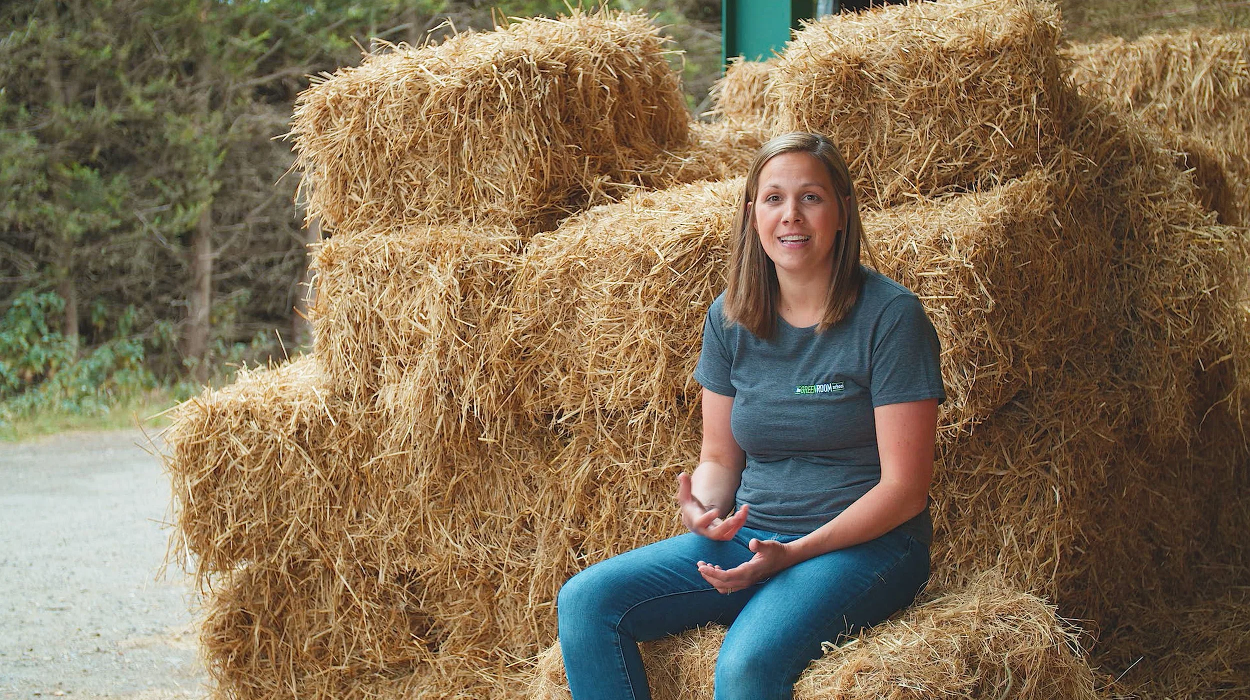 A woman with shoulder-length brown hair sitting on a bale of hay outdoors, holding a white object, with more hay bales behind her and a blurred background of trees.