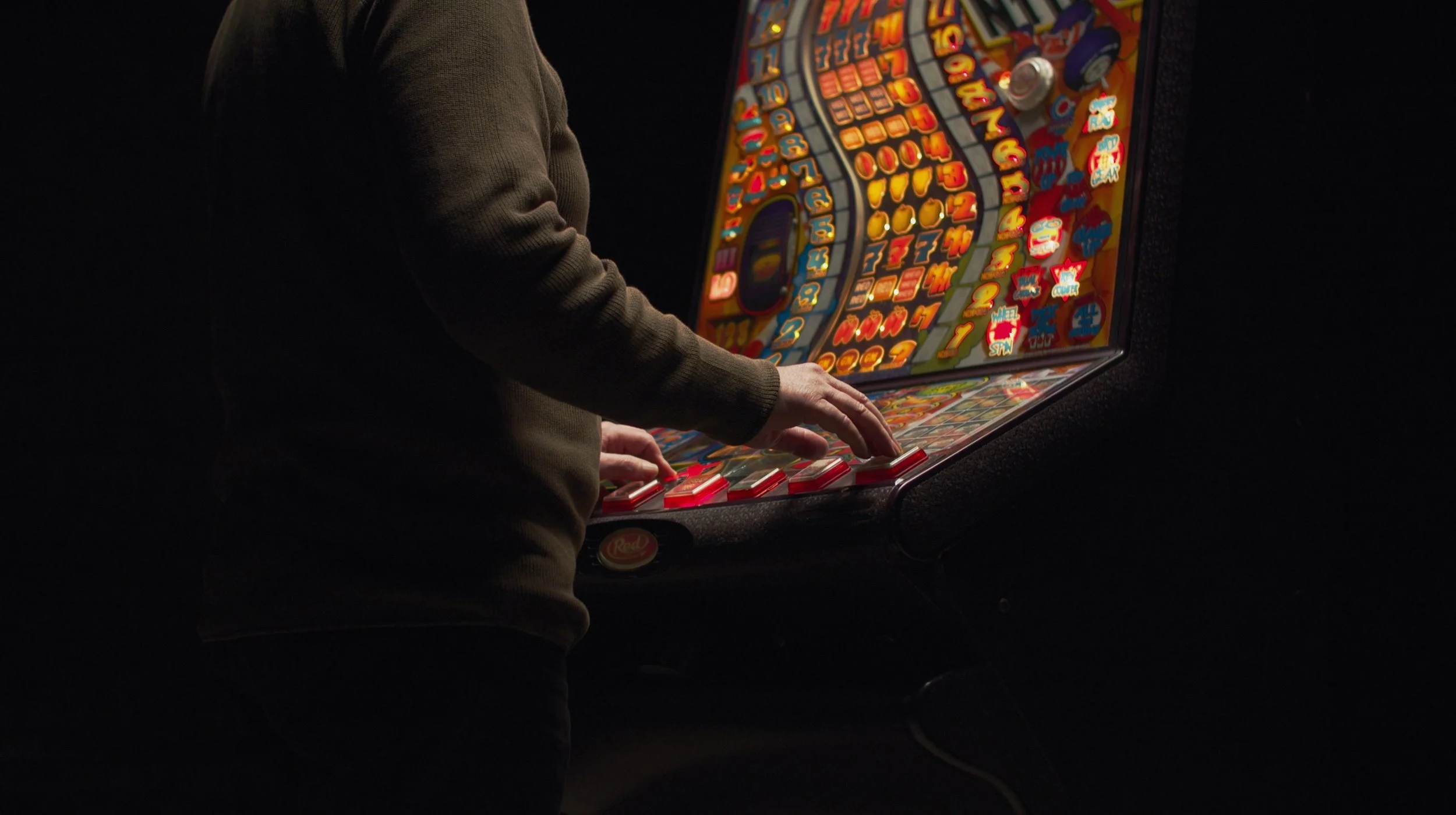 Person playing a colorful pinball or arcade game in a dark room, with their hands on the controls and the game screen illuminated.