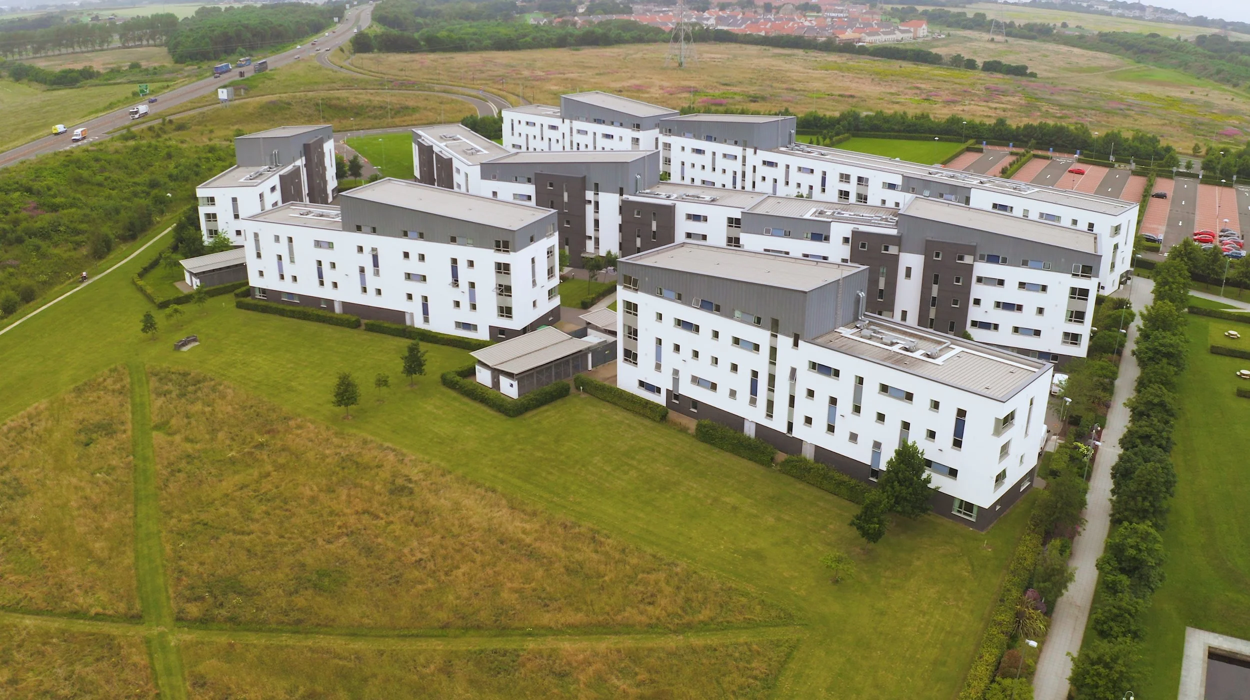 Aerial view of modern white residential buildings with gray accents, trees, paved walkways, parking lots with cars, adjacent grassy fields, highway with vehicles, and distant hills with houses and greenery.