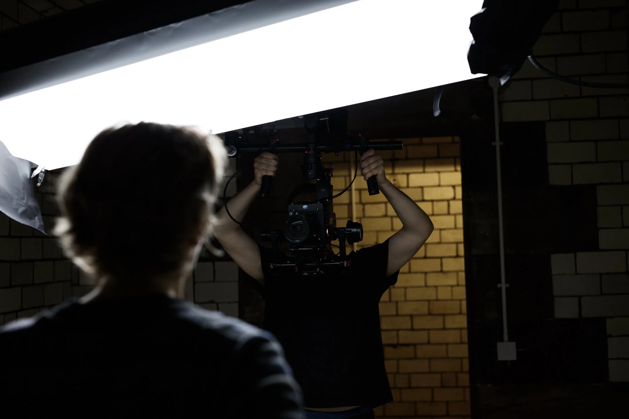 Person adjusting a professional camera setup under bright lighting, with a person observing in the foreground, against a background of brick walls.