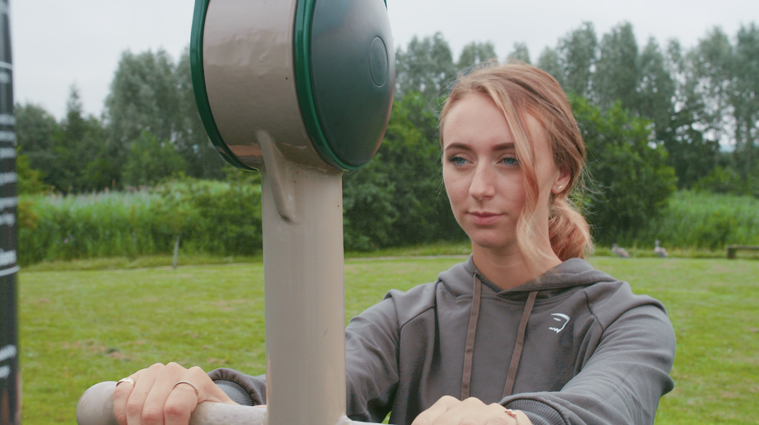 Young woman with red hair exercising outdoors by stretching with a water fountain in the background.