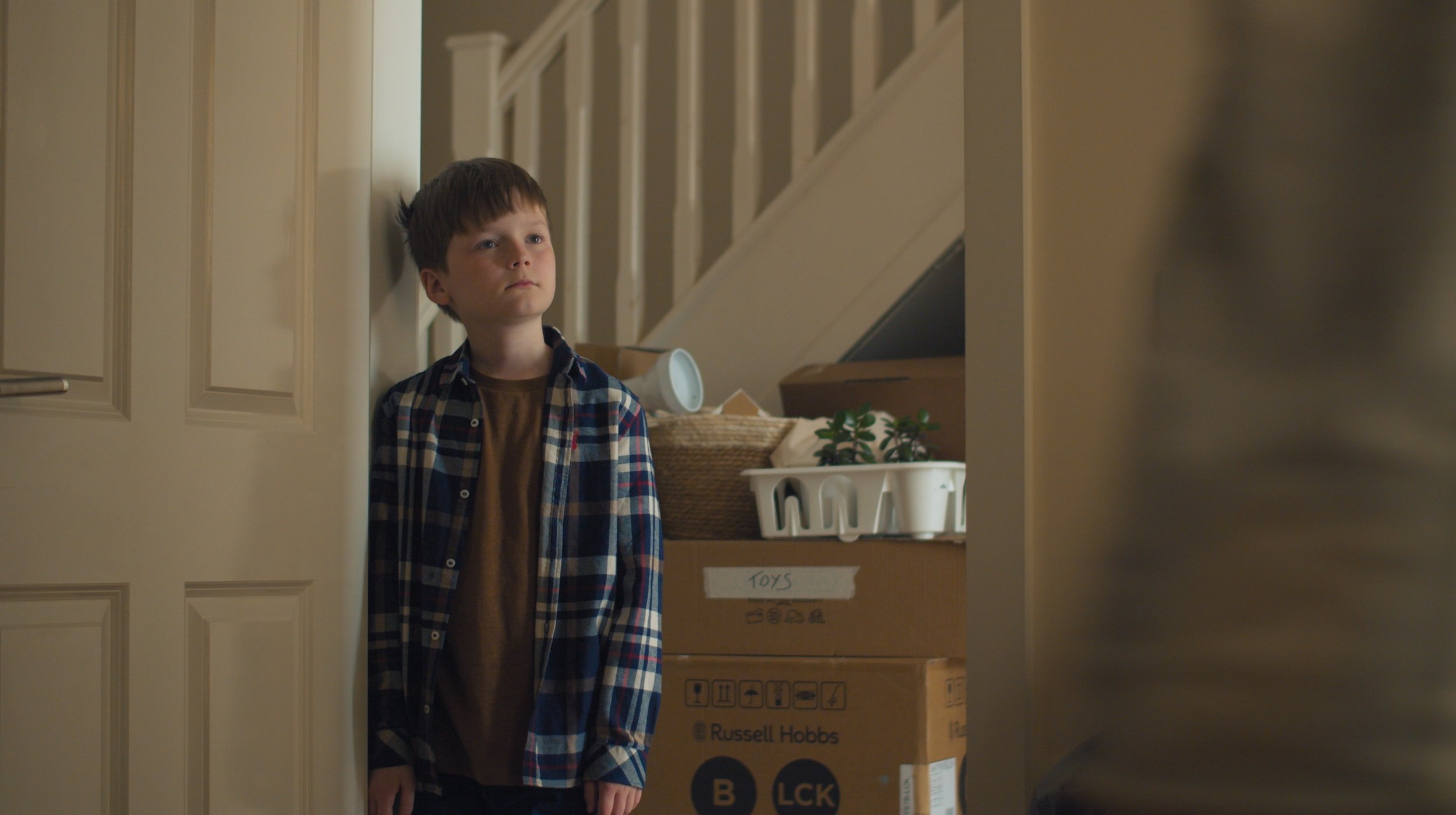 A young boy with brown hair and wearing a plaid shirt and brown T-shirt, standing in a doorway, looking sad or contemplative. Behind him are boxes and storage bins in a home setting.