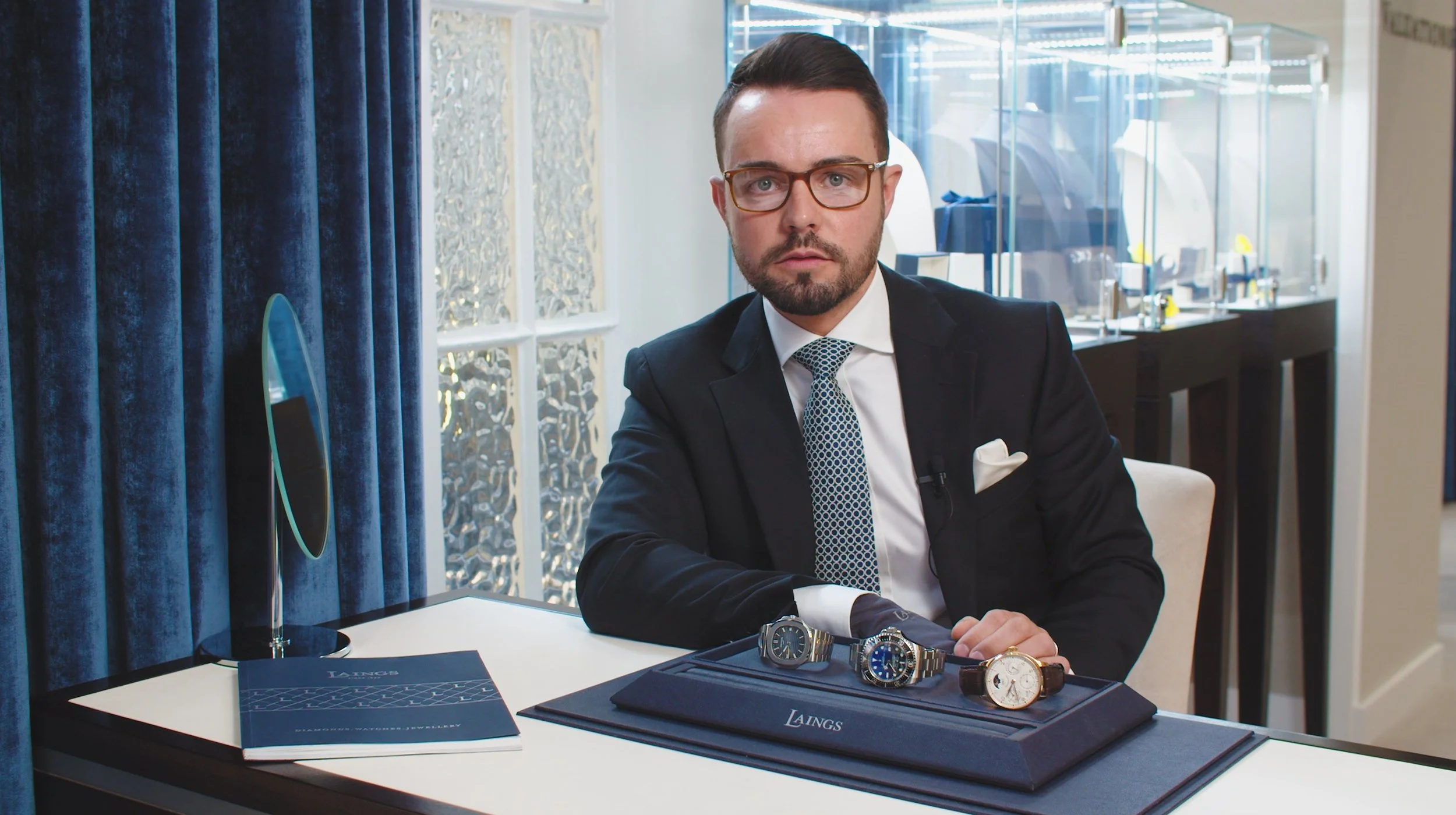 Man in a suit with glasses sitting at a table displaying three luxury watches in a watch case.