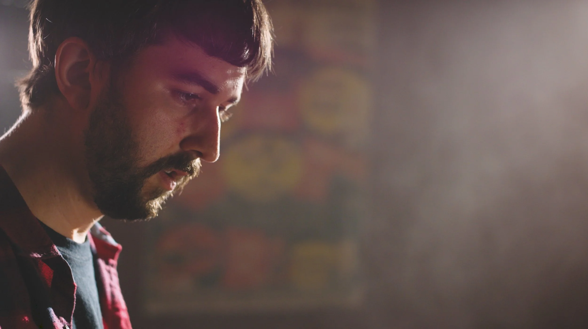 A man with a beard and mustache looking down, in profile, with a blurred colorful background.