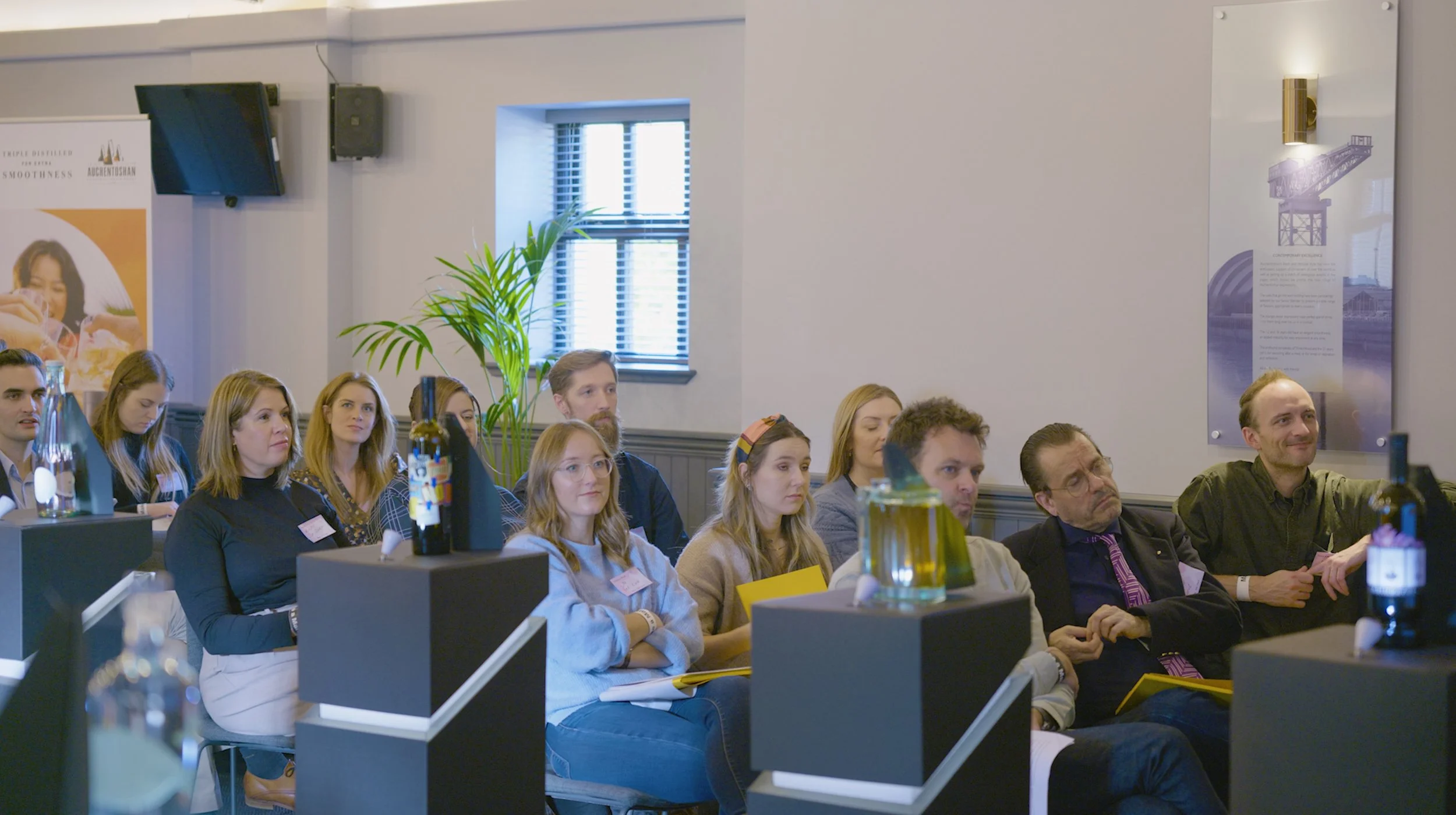 People attending a presentation in a conference room, seated and listening, with bottles and jars on display in the foreground.