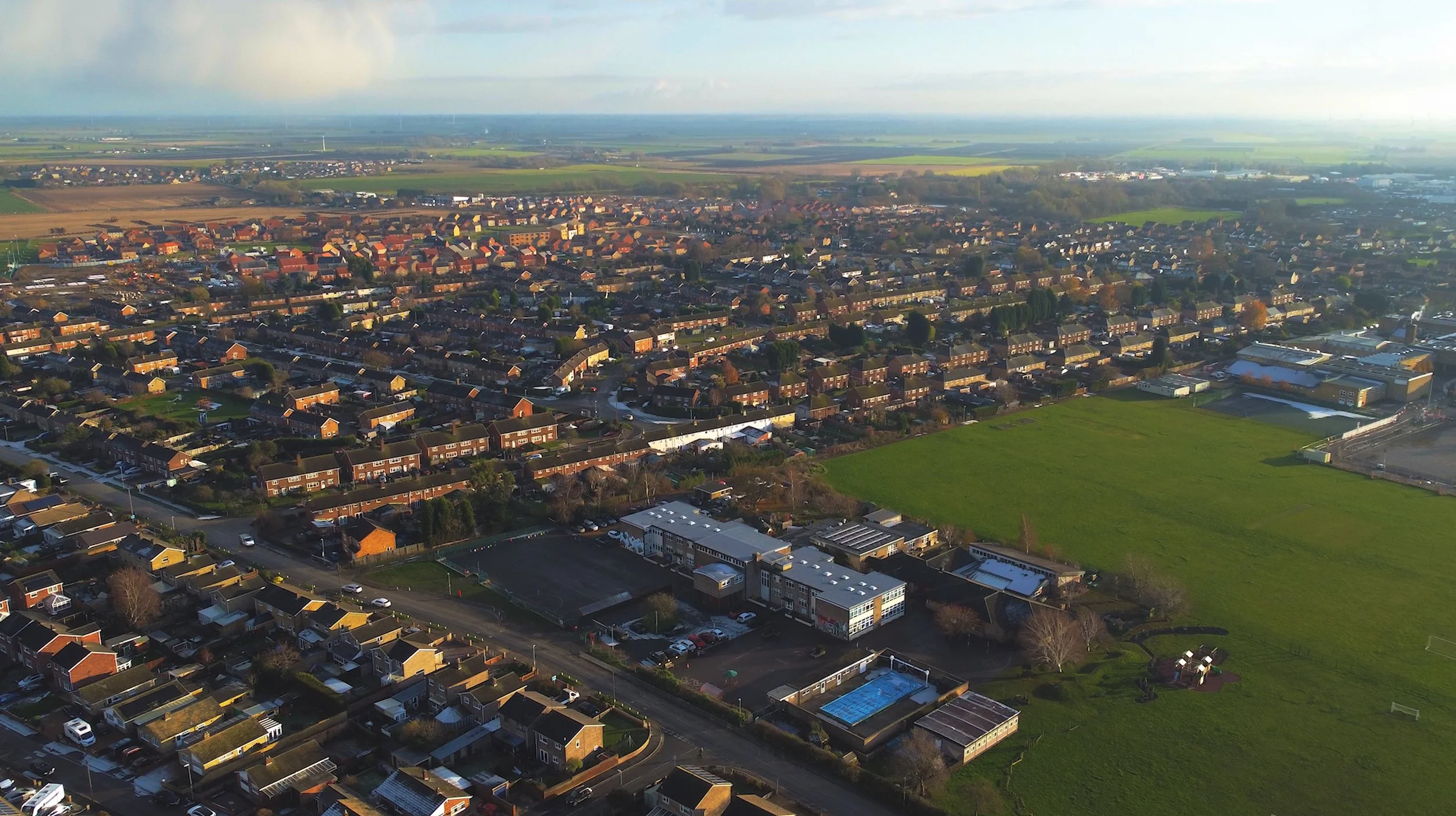 Aerial view of a suburban area with houses, streets, and green fields under a partly cloudy sky.