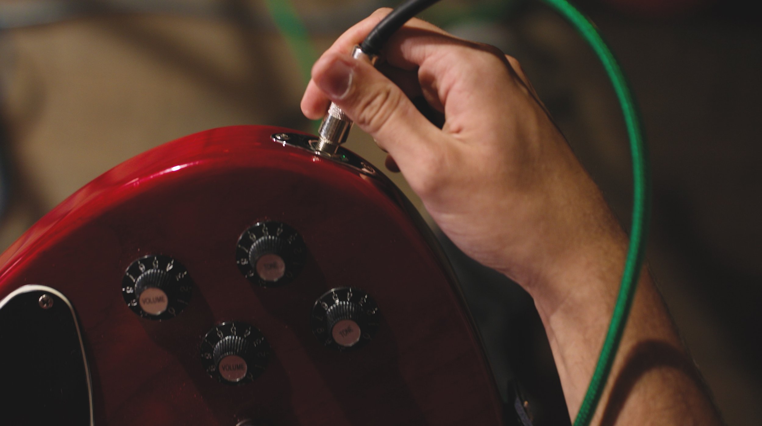 A person adjusting the settings on a red electric guitar using a knob with their right hand, with a green cable plugged into the guitar.