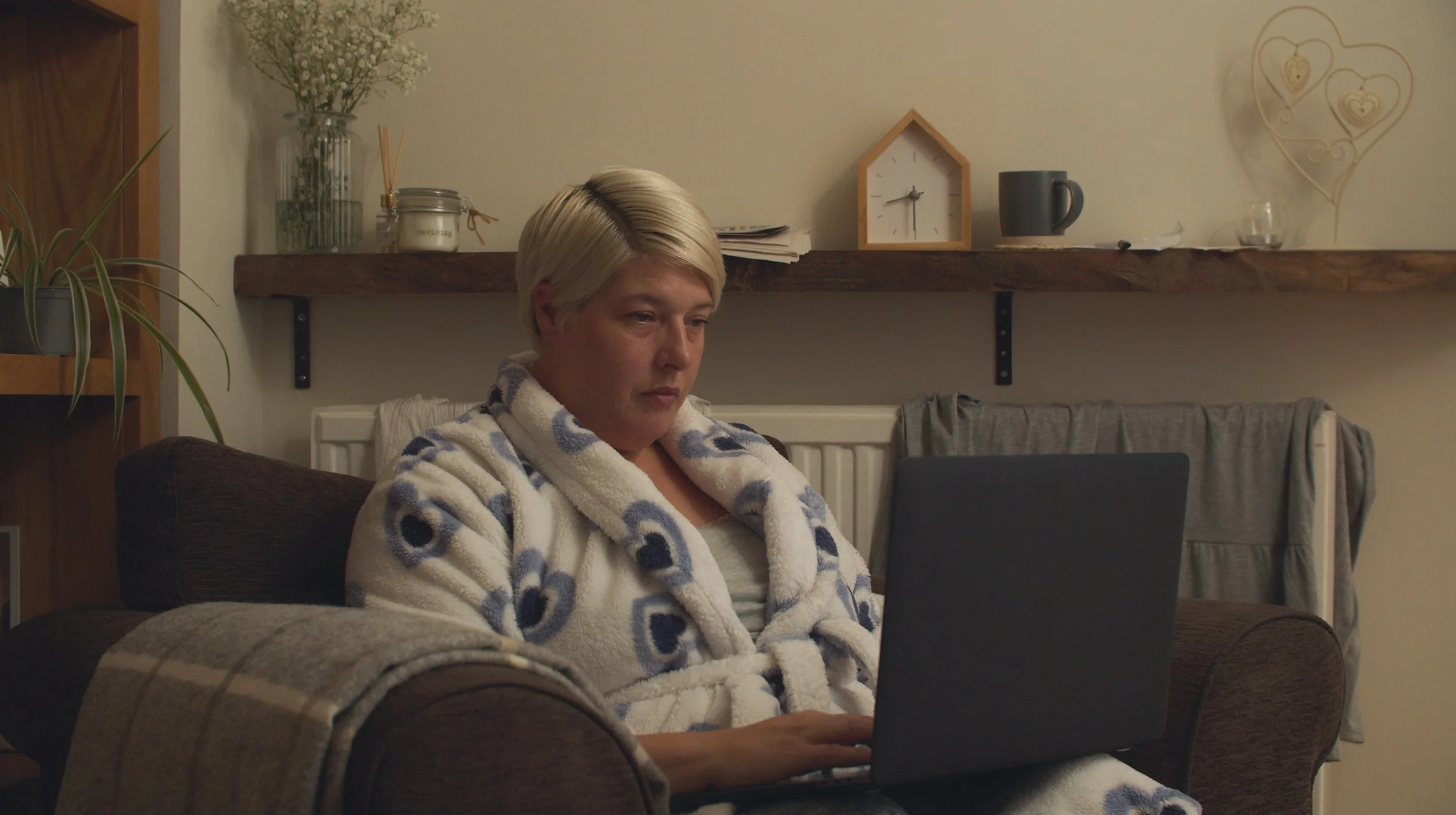 A woman with short blonde hair sitting on a brown couch, wearing a white robe with blue patterns, working on a laptop.