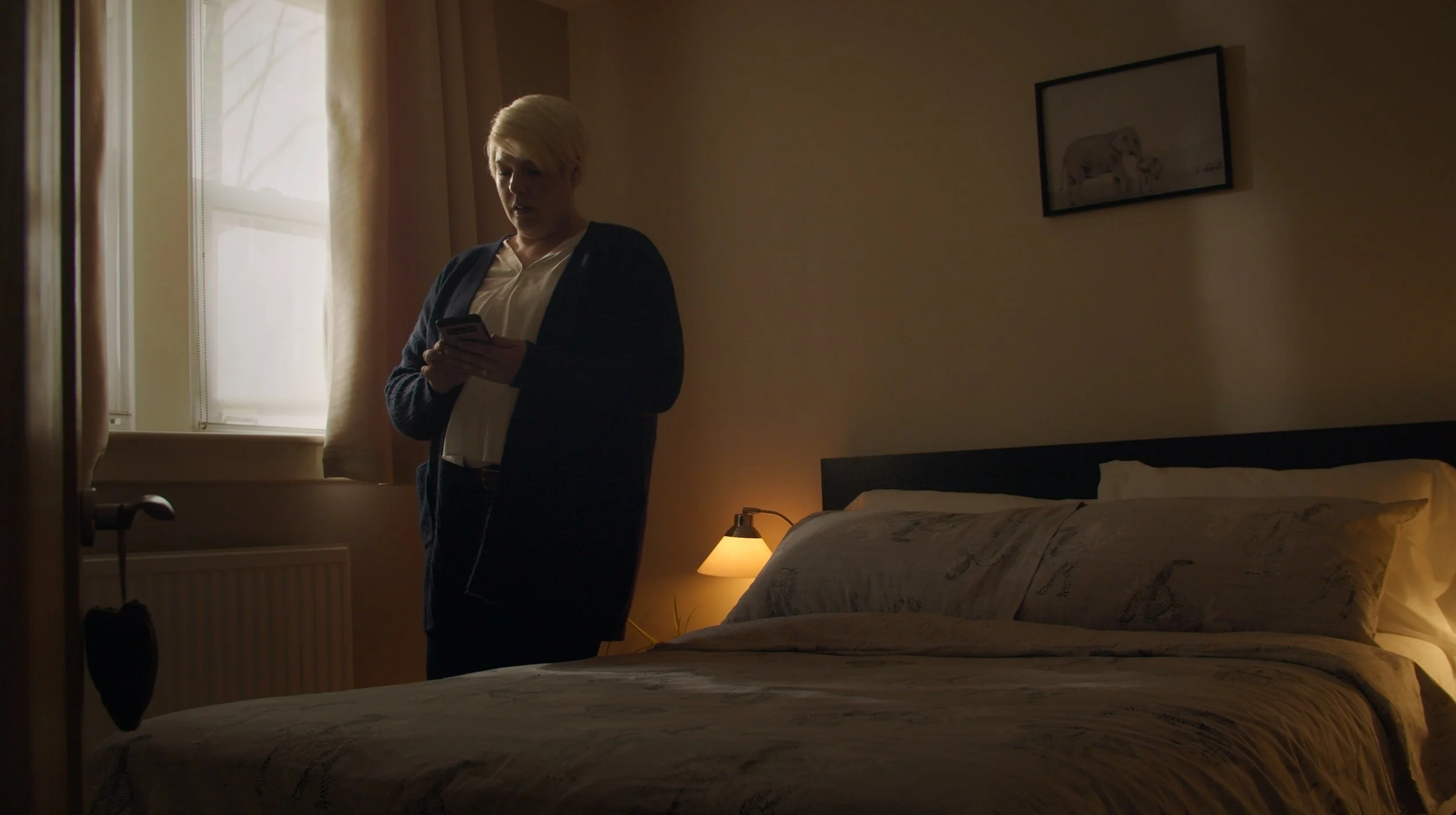 A woman with short blonde hair standing in a dimly lit bedroom, looking at her phone. She is near a window with beige curtains, and a bed with a patterned bedspread. There is a small bedside lamp turned on and a framed picture of an elephant on the wall.