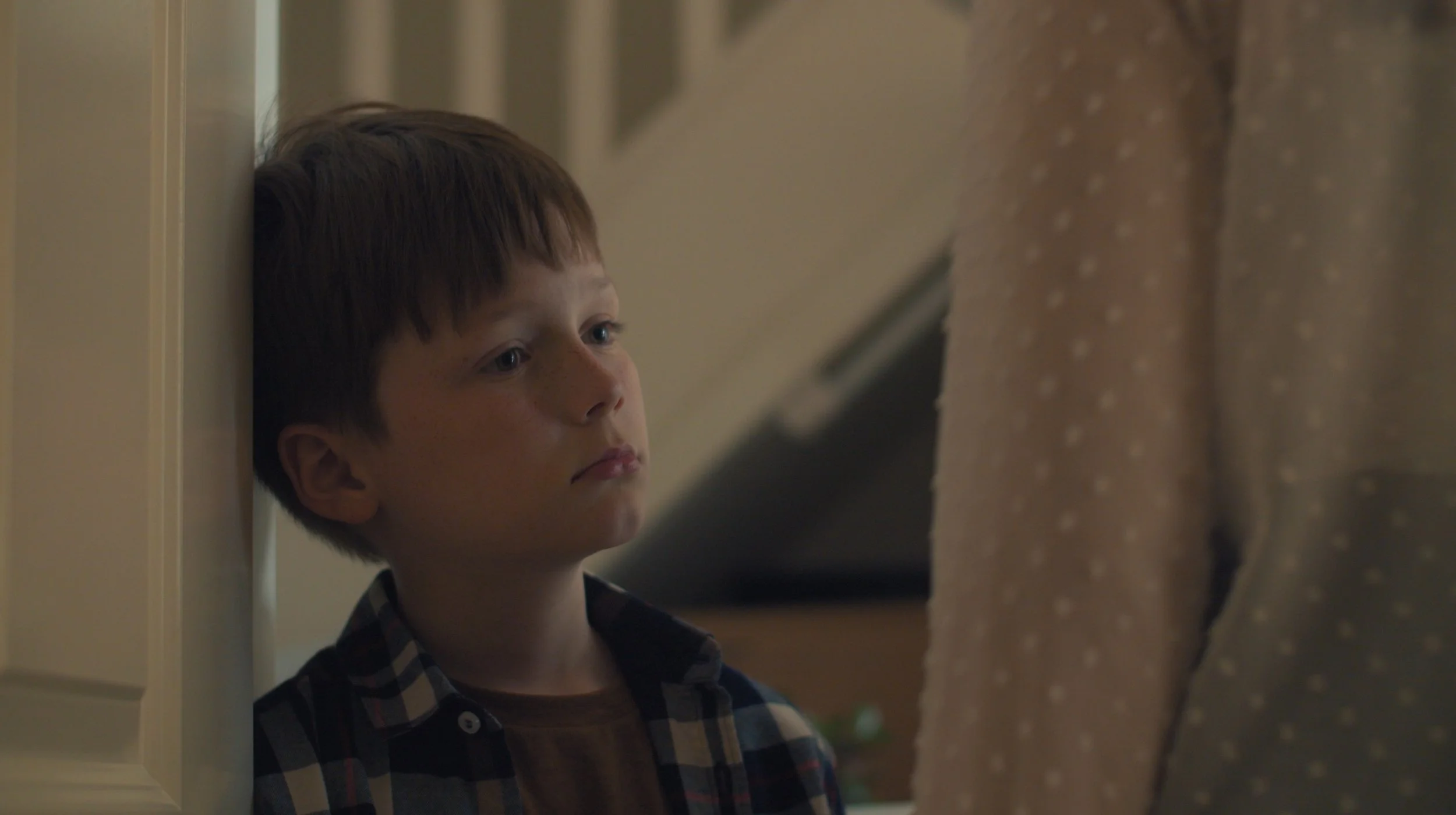 A young boy with brown hair and a checked shirt, looking sad and leaning against a wall, with an adult standing nearby.