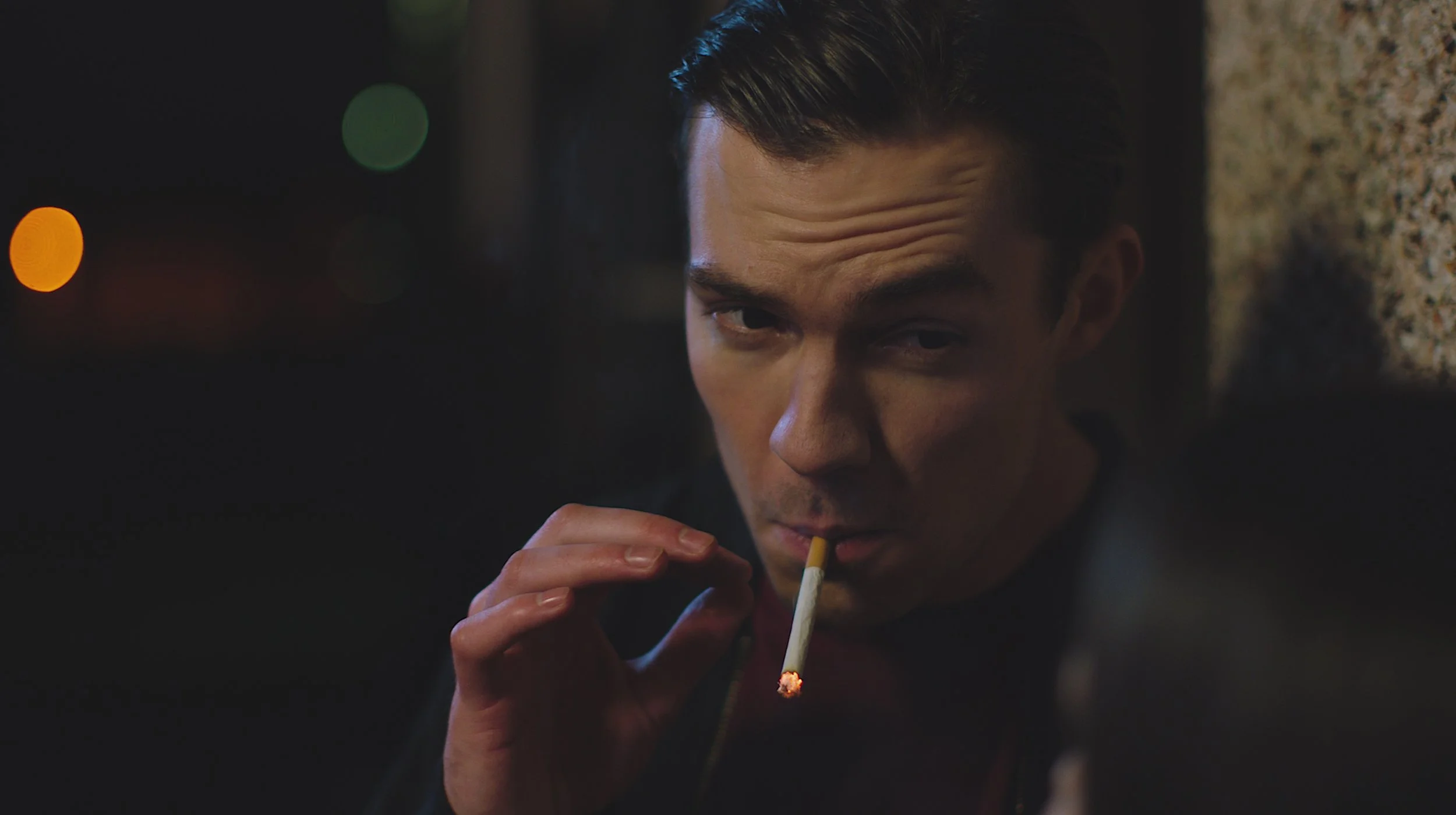 A young man with dark hair smoking a cigarette at night, with colorful blurred lights in the background.