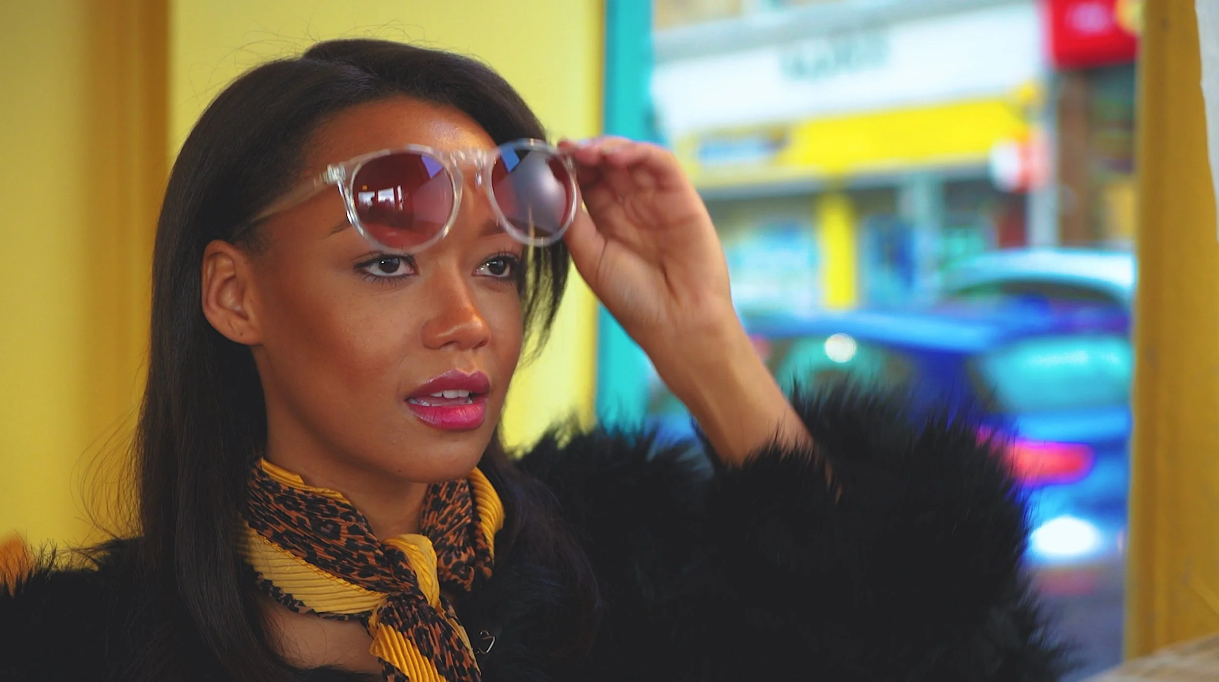 A woman with medium skin tone wearing sunglasses on her head, a yellow and black scarf, and a black fuzzy coat, standing in front of a colorful, blurred background with yellow, blue, and red hues.