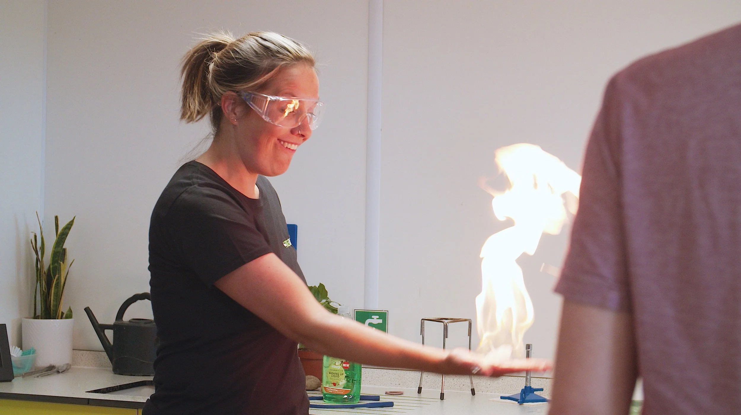 A woman wearing safety goggles, smiling, as she holds her hand that is engulfed in flames in a science lab.