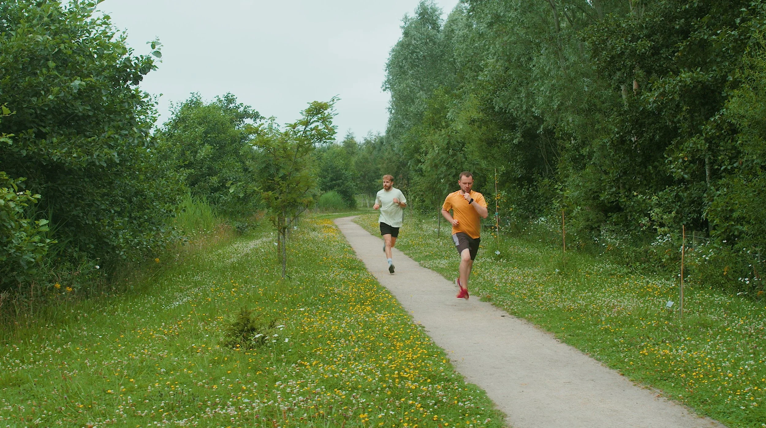 Two men jogging on a narrow paved trail through a lush green park with trees and wildflowers.