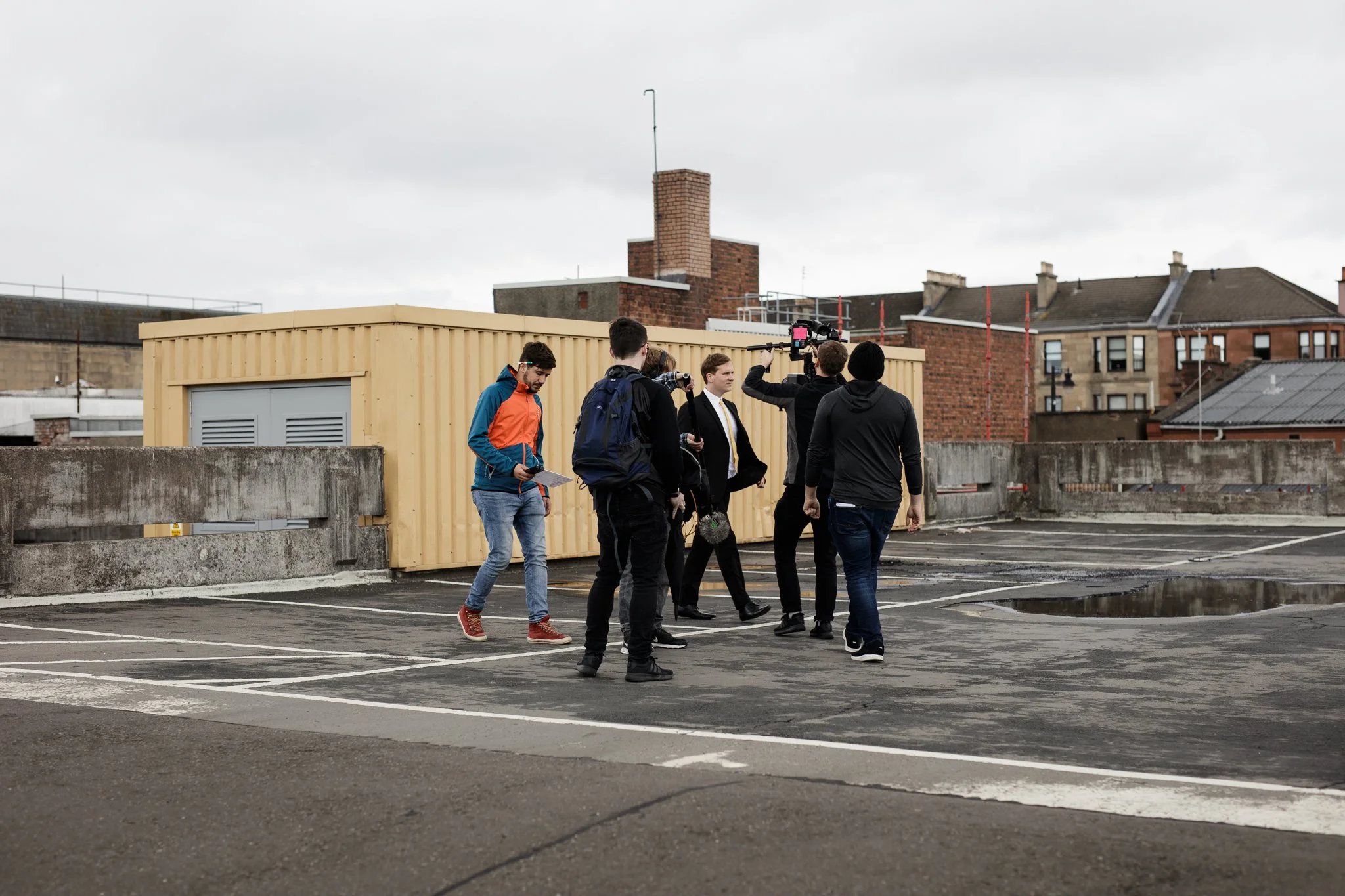 Group of people, including a man in a suit, filming on a rooftop parking lot under cloudy sky, with buildings in the background.