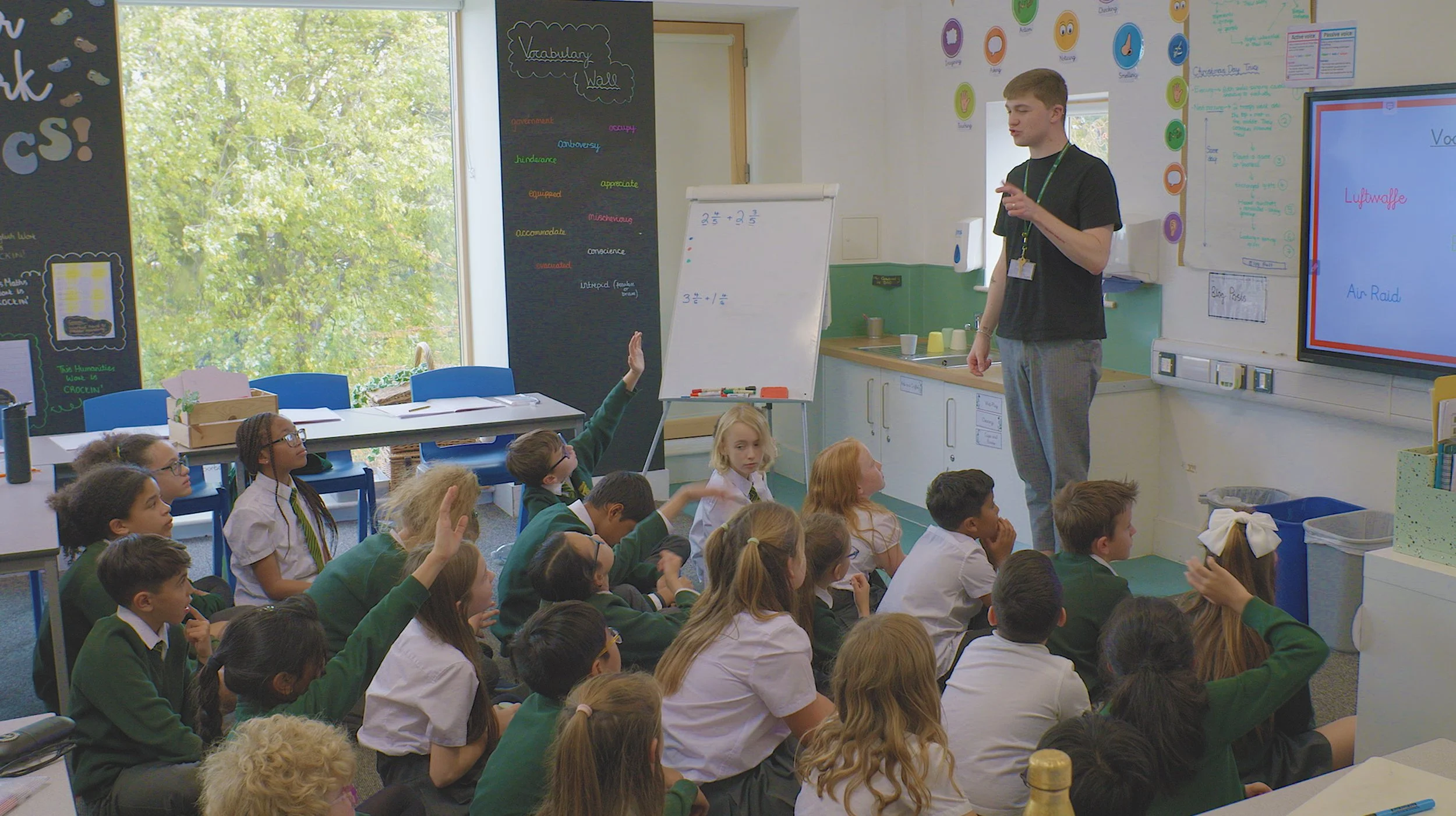 A classroom with students sitting on the floor, raising their hands to ask questions, while a teacher stands beside a whiteboard. The room has colorful posters and a large window with trees outside.