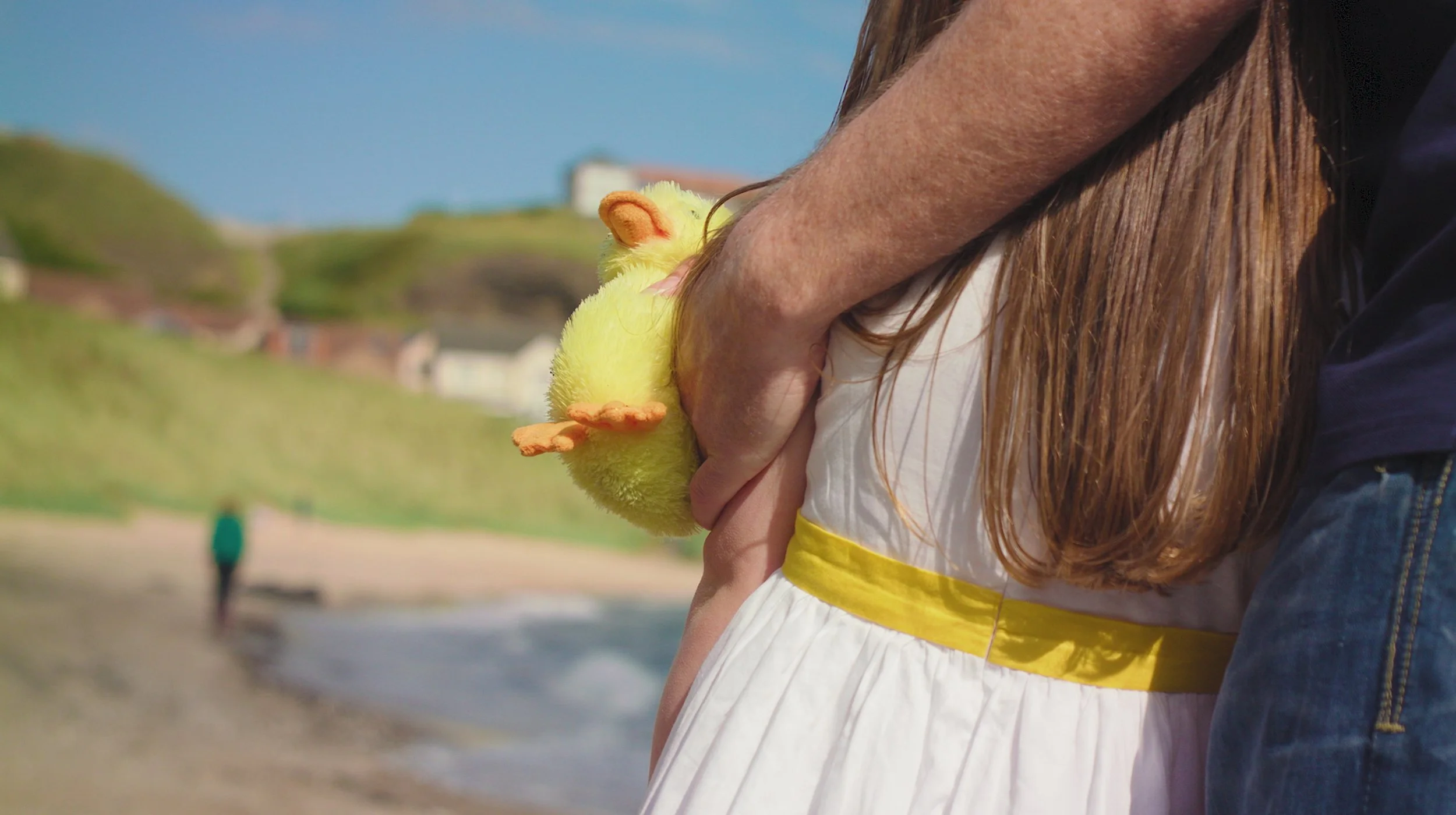 A person holding a yellow stuffed duck toy on a beach, with a grassy hill and houses in the background.