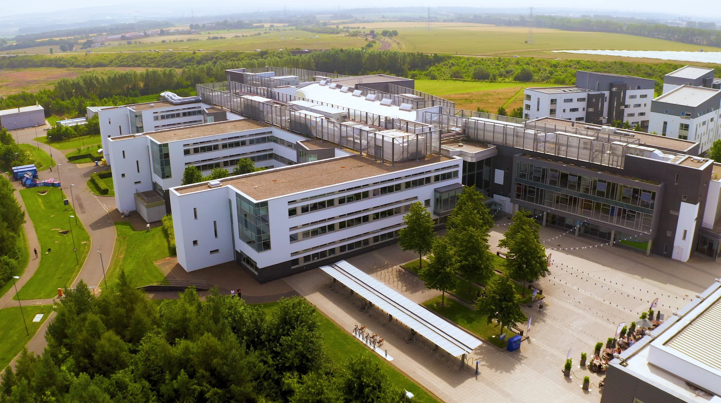 An aerial view of a modern university campus with multiple white and black buildings, green trees, a bicycle parking area, and surrounding open fields.
