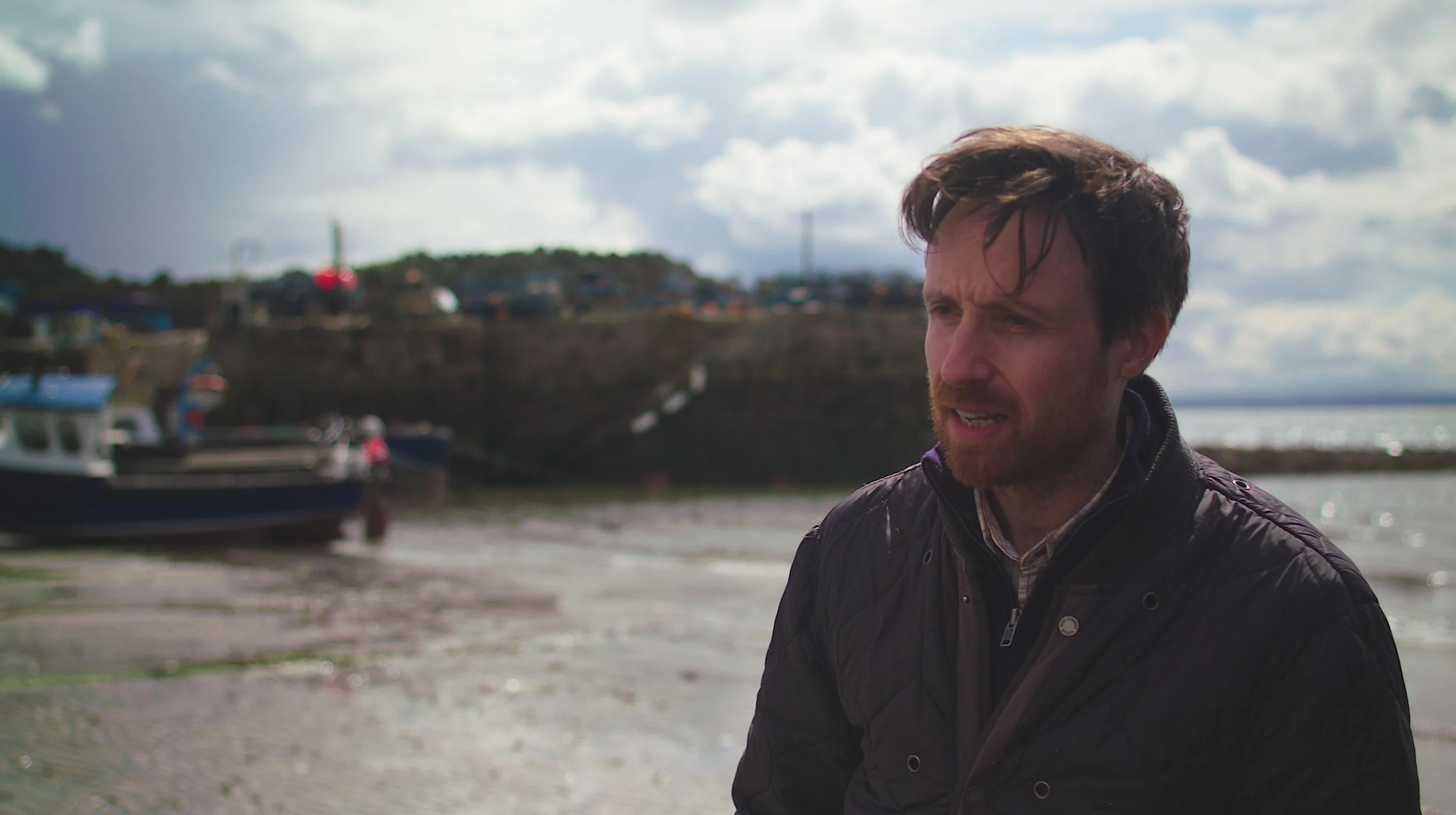 A man with brown hair and a beard standing on a beach near boats, with a cloudy sky in the background.