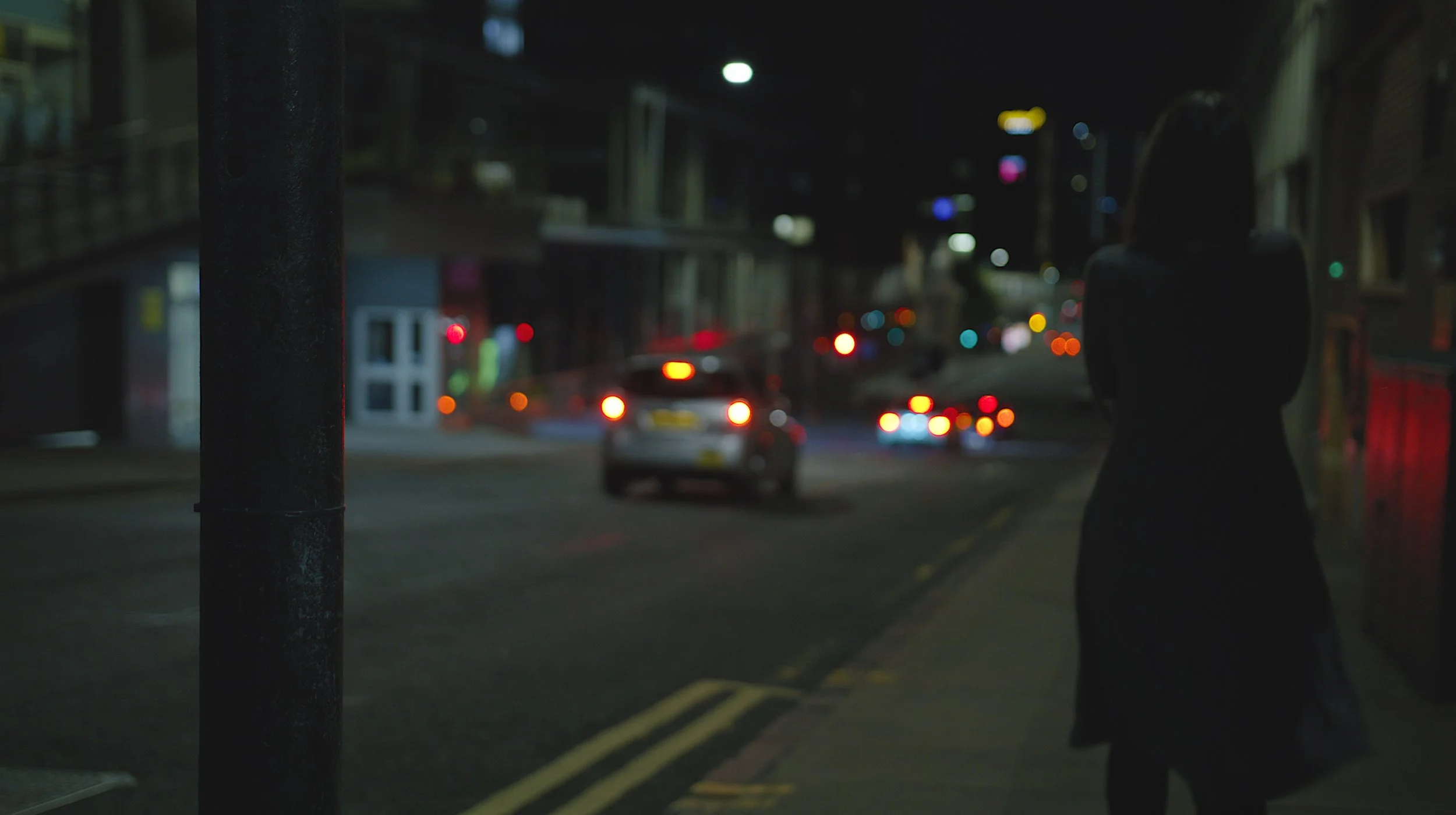 A person dressed in dark clothing walking on a city sidewalk at night, with blurred cars and streetlights in the background.