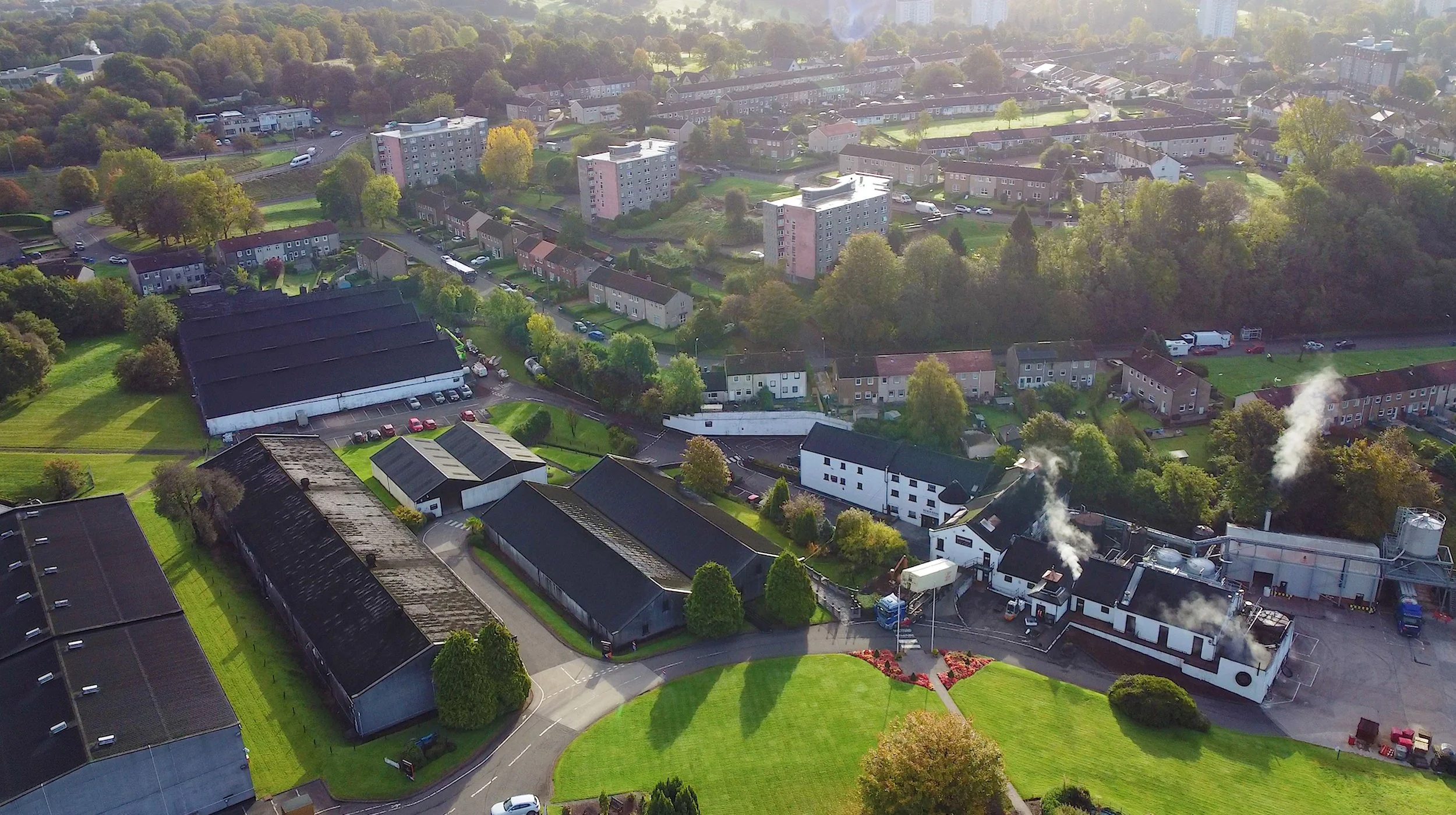Aerial view of an industrial complex with white and black buildings, some emitting white smoke, surrounded by green trees and a residential neighborhood with houses, apartments, and parking lots.