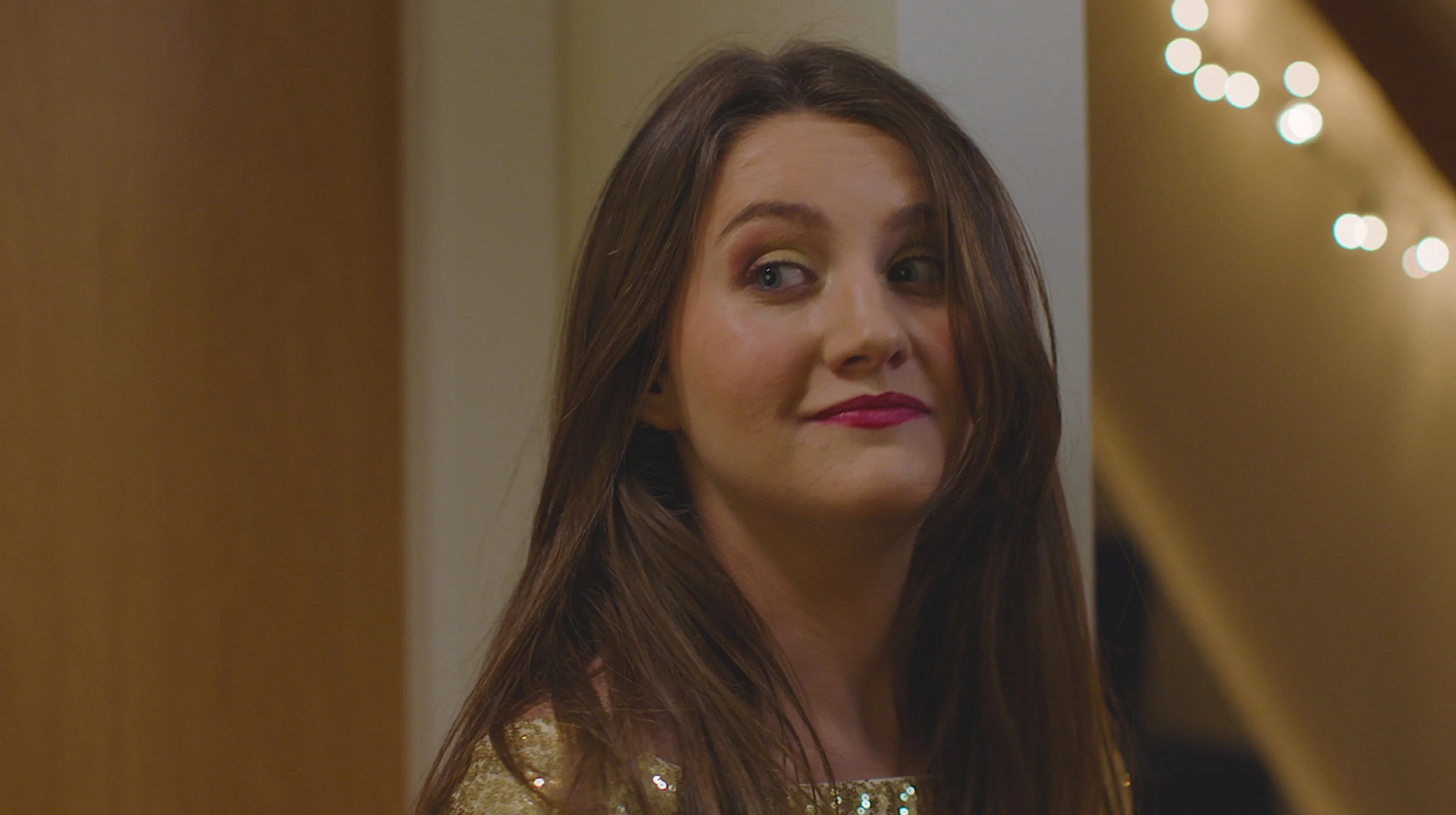 A young woman with long brown hair, wearing a gold sequin dress, looking to her left with a smirk on her face. In the background, there are blurred string lights and a wooden wall.