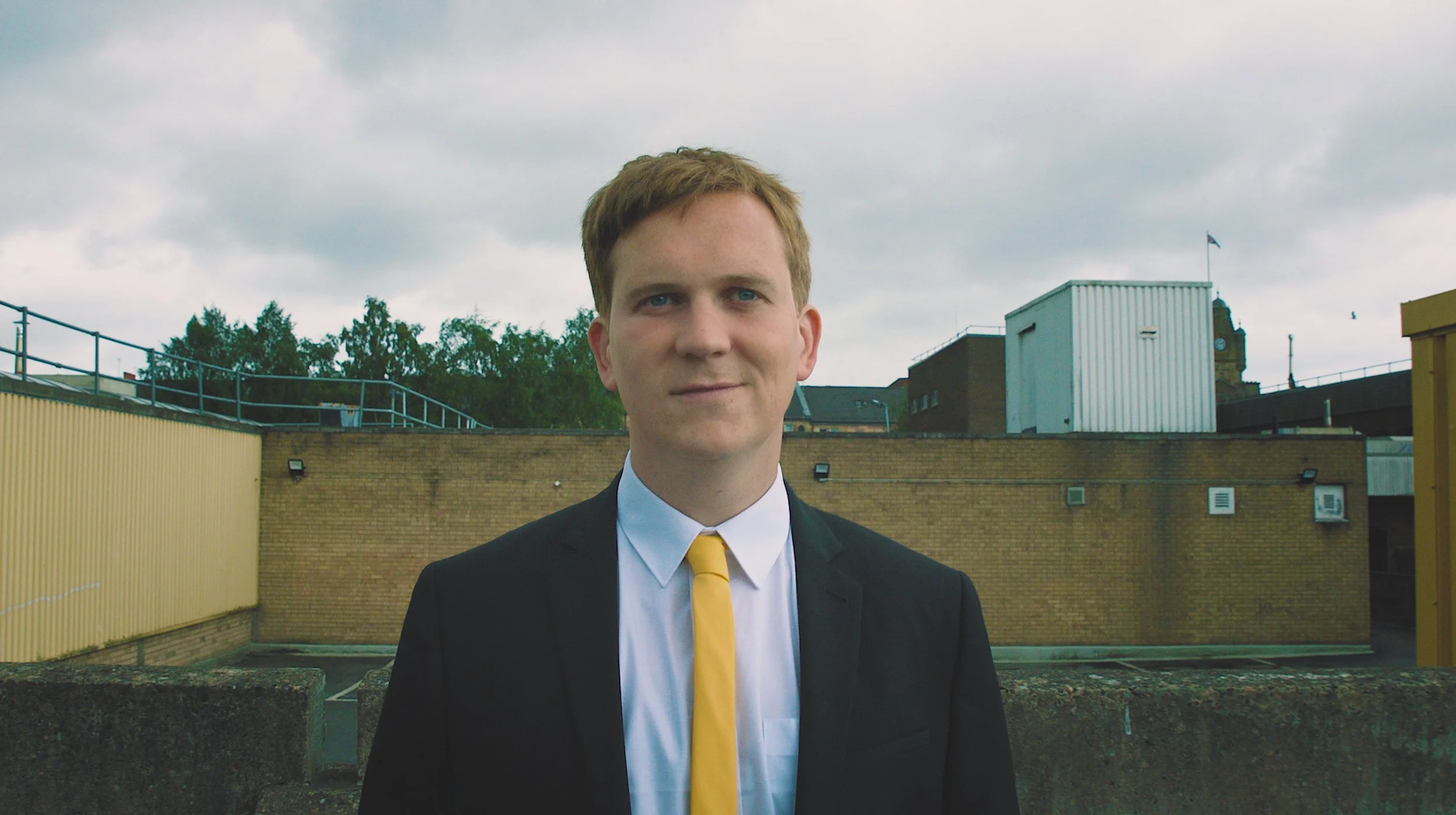 A young man with short reddish hair in formal business attire, including a black suit, white shirt, and yellow tie, standing outdoors on a rooftop with a brick wall and industrial buildings in the background under a cloudy sky.