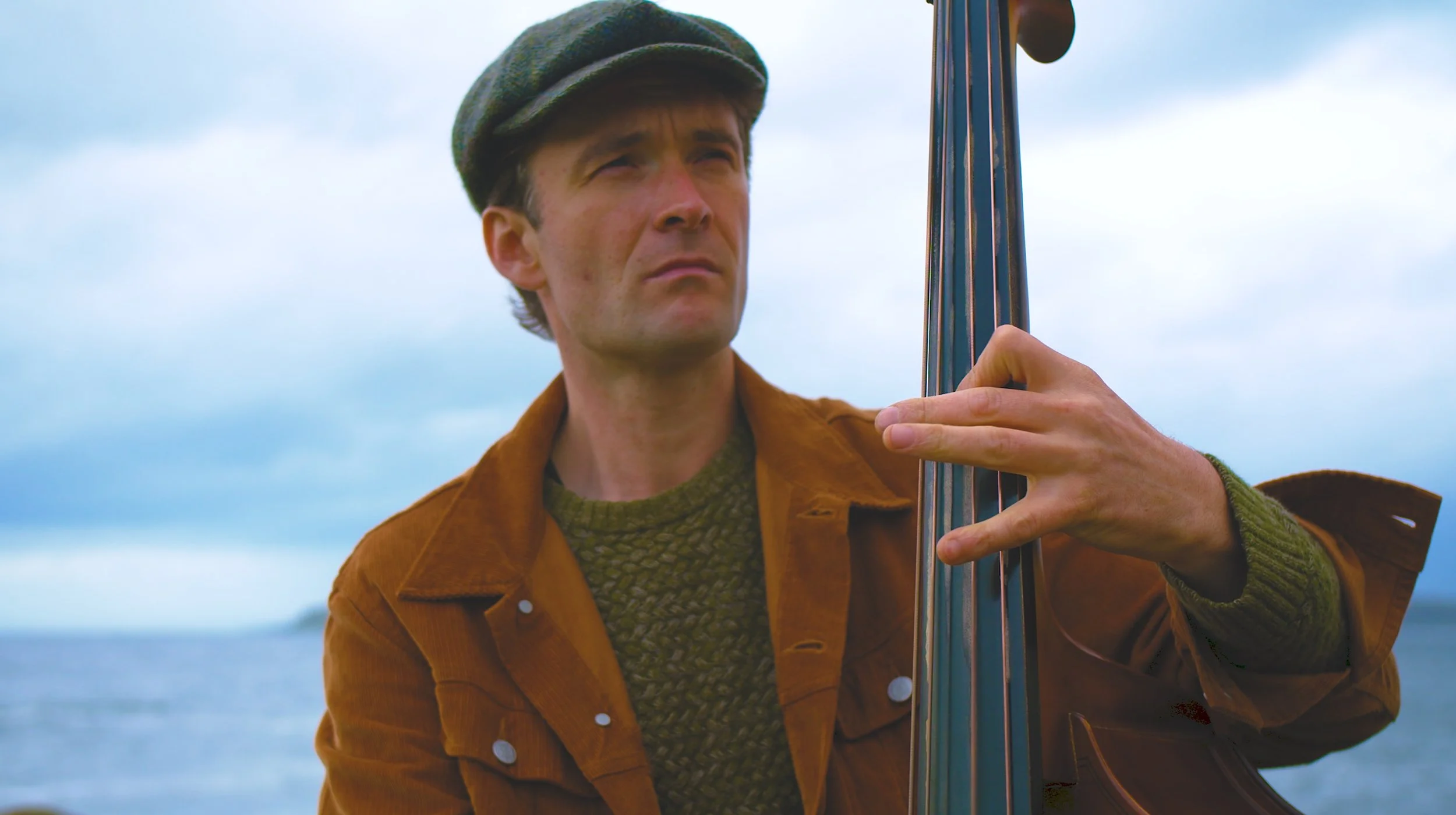 A man wearing a green flat cap and brown jacket playing a double bass outdoors, with a cloudy sky and body of water in the background.