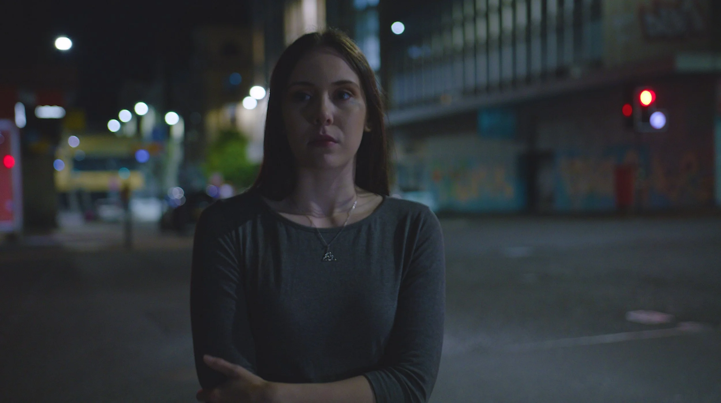 A young woman with red hair stands on a city street at night, crossing her arms and looking to the side, illuminated by streetlights and traffic signals.